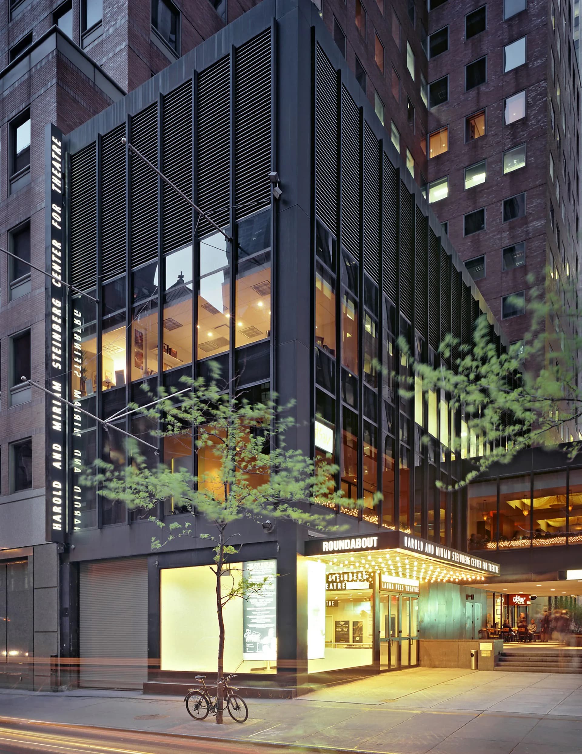 Modern urban ground-floor retail and office building with dark metal lattice screening, illuminated interior spaces, and verdant planted areas visible through large windows, designed by Fred Basch Architect PLLC.