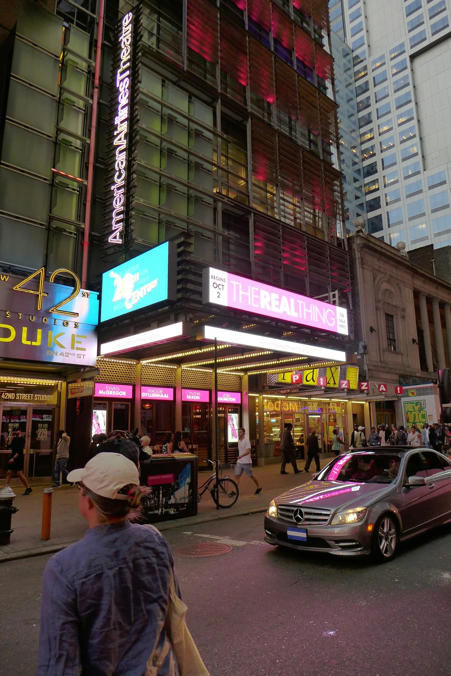 Urban street-level storefront with illuminated signage featuring pink and cyan neon lighting, multi-story facade with modern digital displays, surrounded by tall buildings in a dense city environment by Fred Basch Architect PLLC.