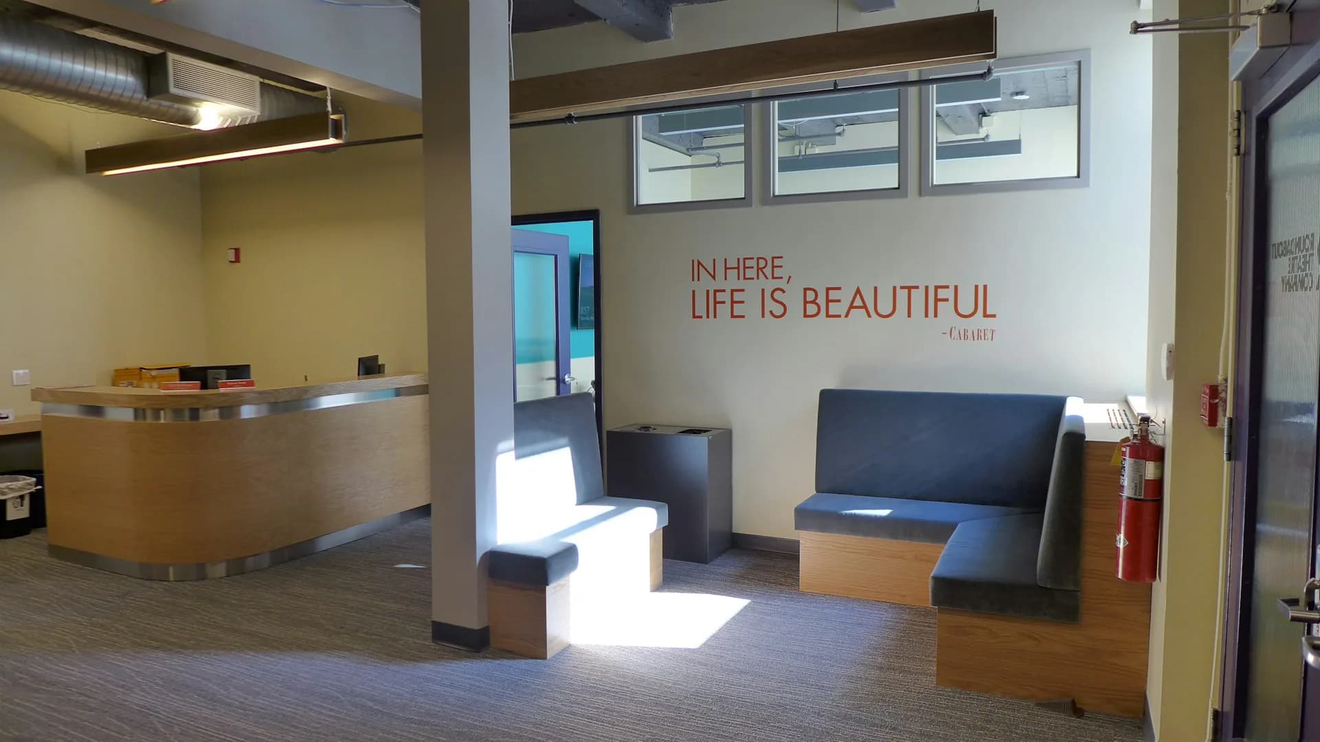 Modern office reception area featuring a curved wooden desk, blue seating, concrete floors, and inspirational wall text, designed by Fred Basch Architect PLLC.