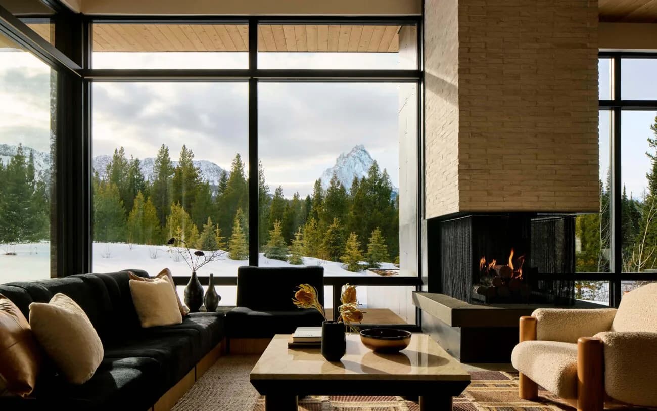 Modern mountain lodge interior with black fireplace, dark sofa, and expansive windows framing snow-capped peaks and forest by Frederick Tang Architecture.