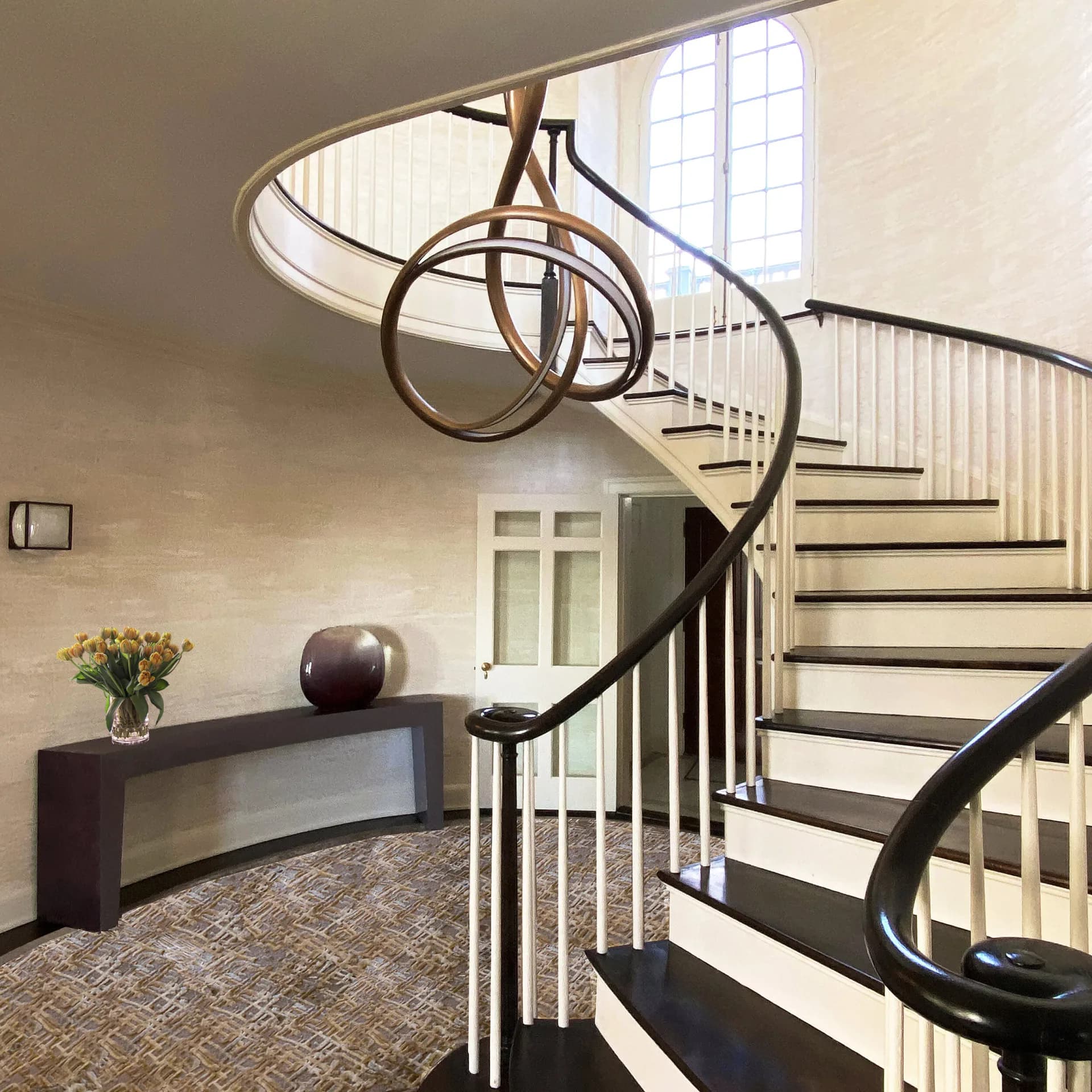 Modern spiral staircase with black metal railings and curved sculptural element in cream-toned foyer with dark console table by Fresco Decorative Arts.