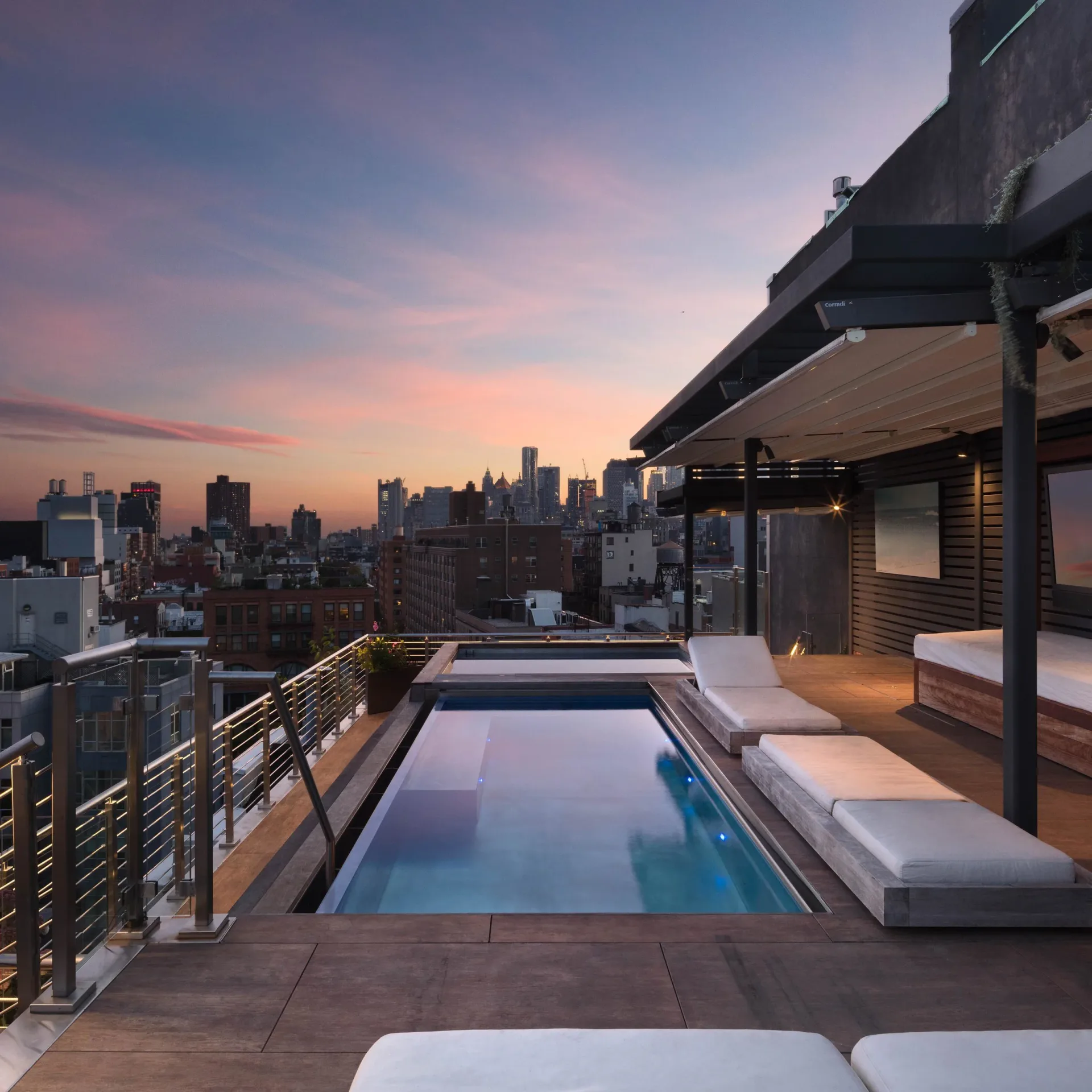Rooftop pool deck at dusk overlooking Manhattan skyline, featuring a narrow lap pool, lounge seating, pergola structure, and metal railing by Freyer Collaborative Architects.