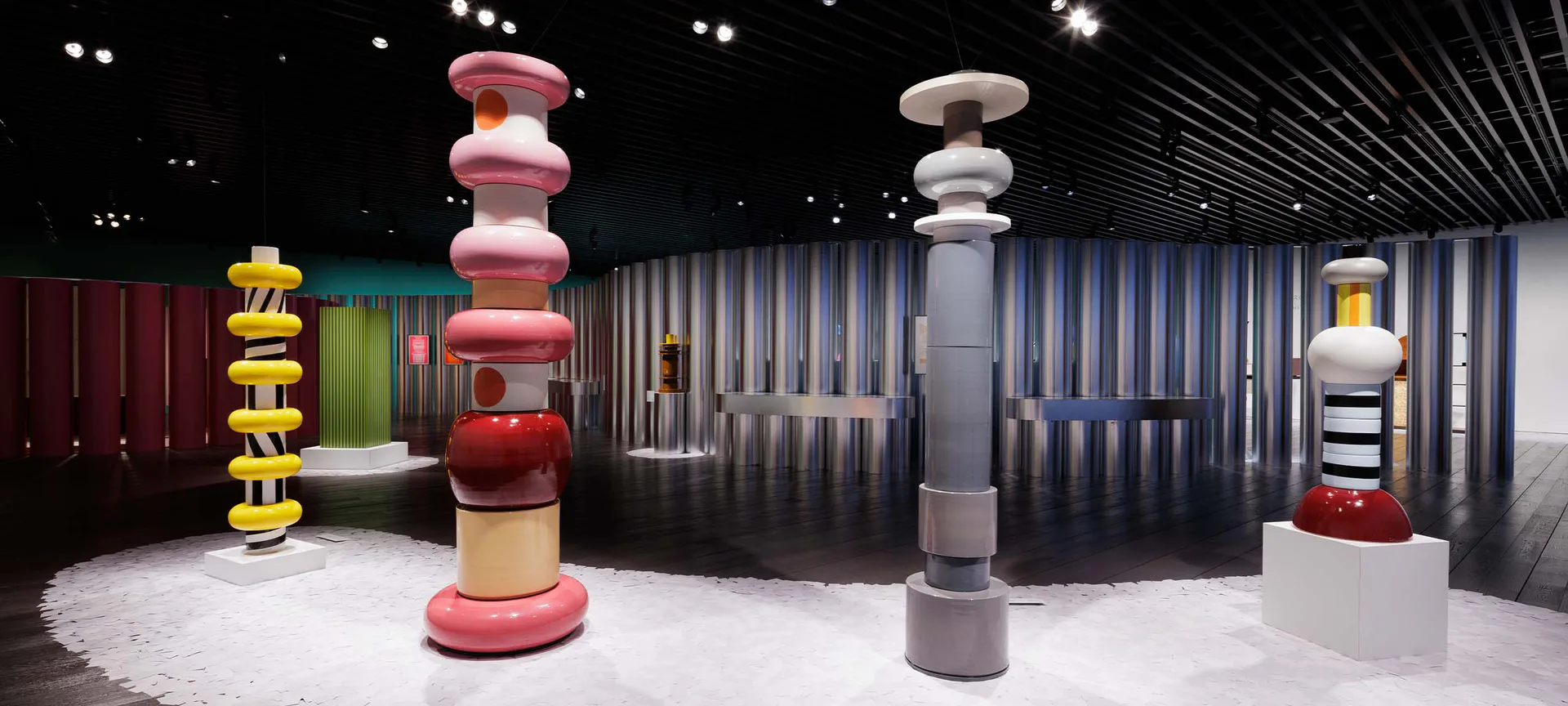 Abstract exhibition space with stacked sculptural forms in red, gray, and yellow tones arranged on white flooring, geometric metal wall paneling, and dramatic lighting by Friedman Benda.