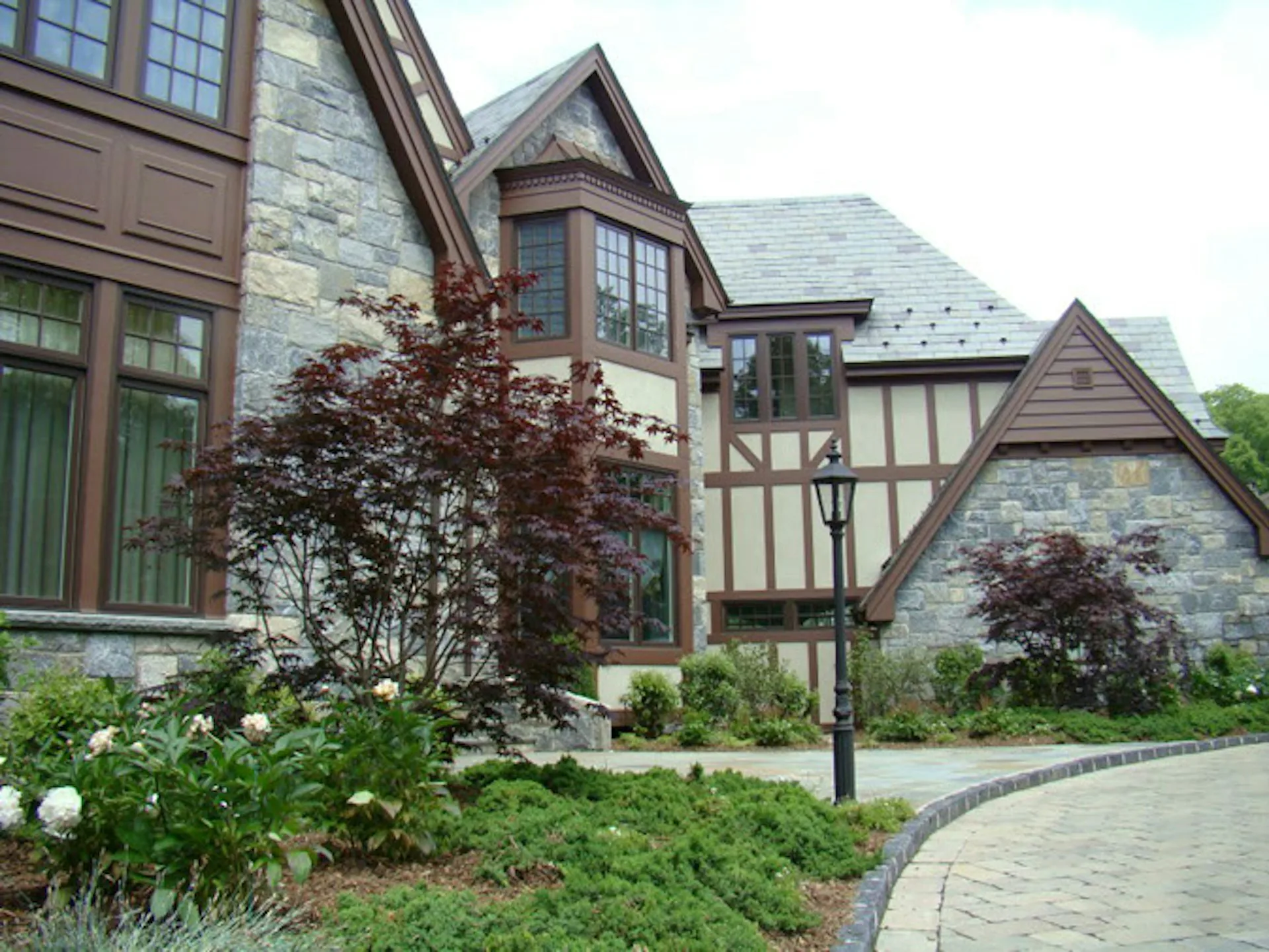 Tudor-style residential exterior featuring stone and timber detailing, manicured landscaping with ornamental shrubs, and curved stone driveway by FSI Architecture PC.