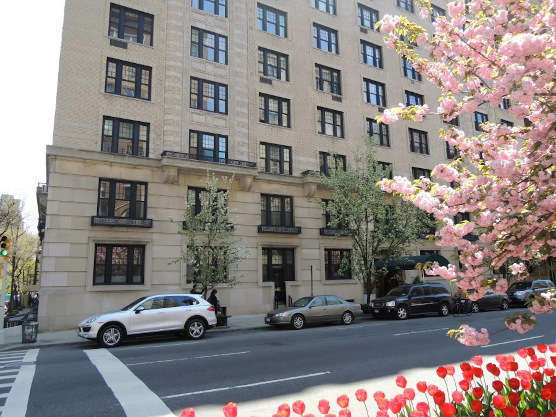 Street-level facade of a stone residential building with regular window arrangement, parked vehicles, and blooming pink flowering trees by FSI Architecture PC.