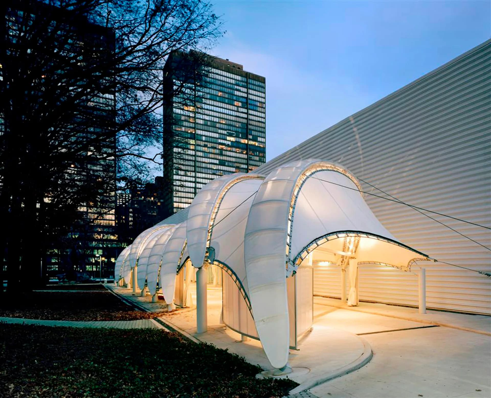 Illuminated white tensile fabric pavilion with curved canopy design at dusk, situated in urban landscape with modern highrise building in background, by FTL DESIGN ENGINEERING STUDIO.