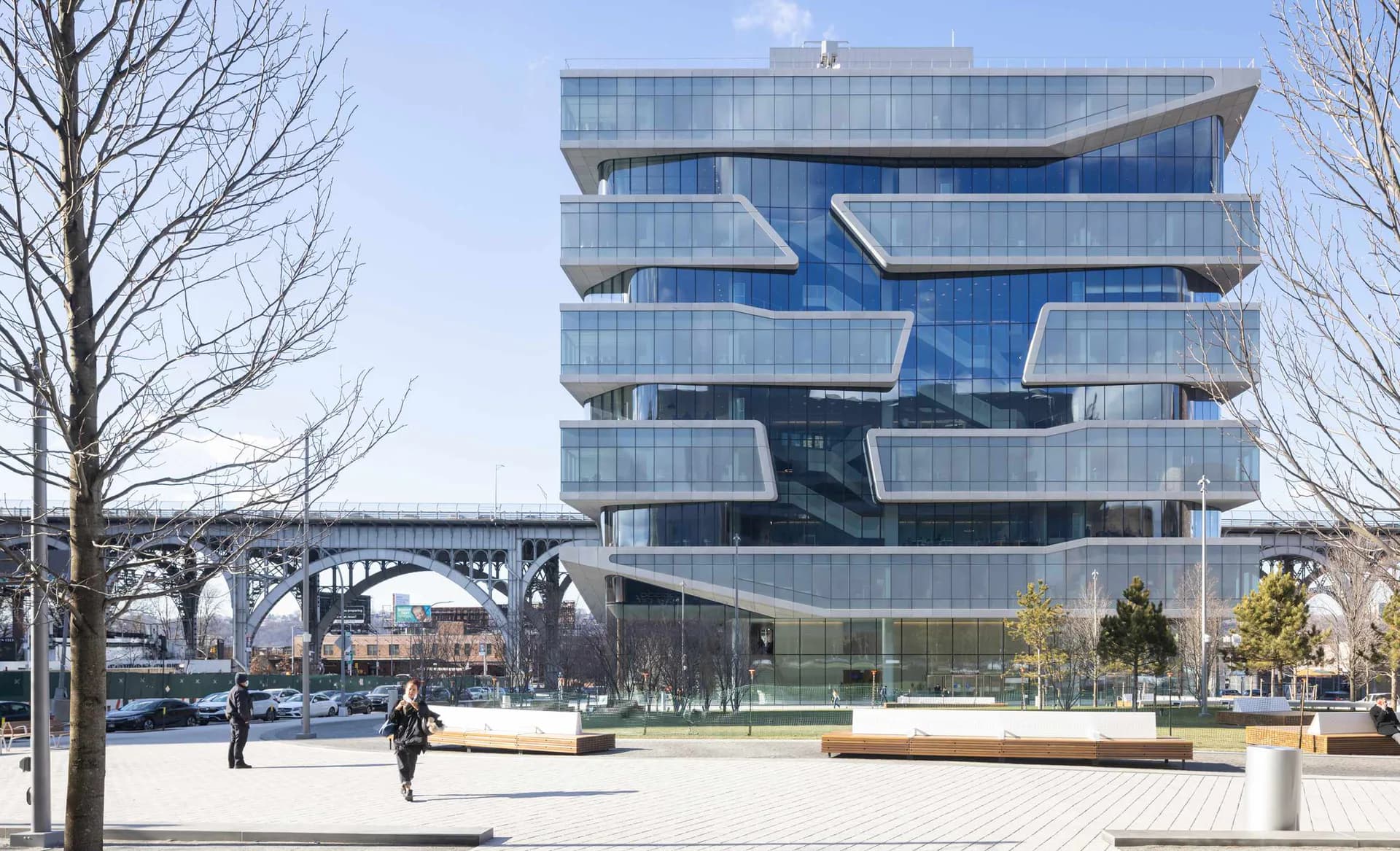 Modern multi-story glass and blue facade building with staggered balconies overlooking a plaza with bare trees and people, by FXCollaborative.