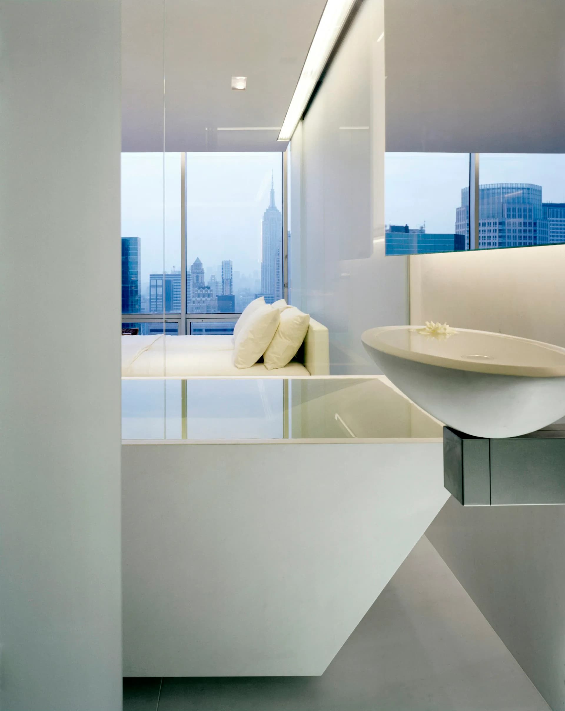 Minimalist bathroom with white fixtures and cabinetry, floor-to-ceiling windows framing Manhattan skyline views, by Gabellini Sheppard Associates.