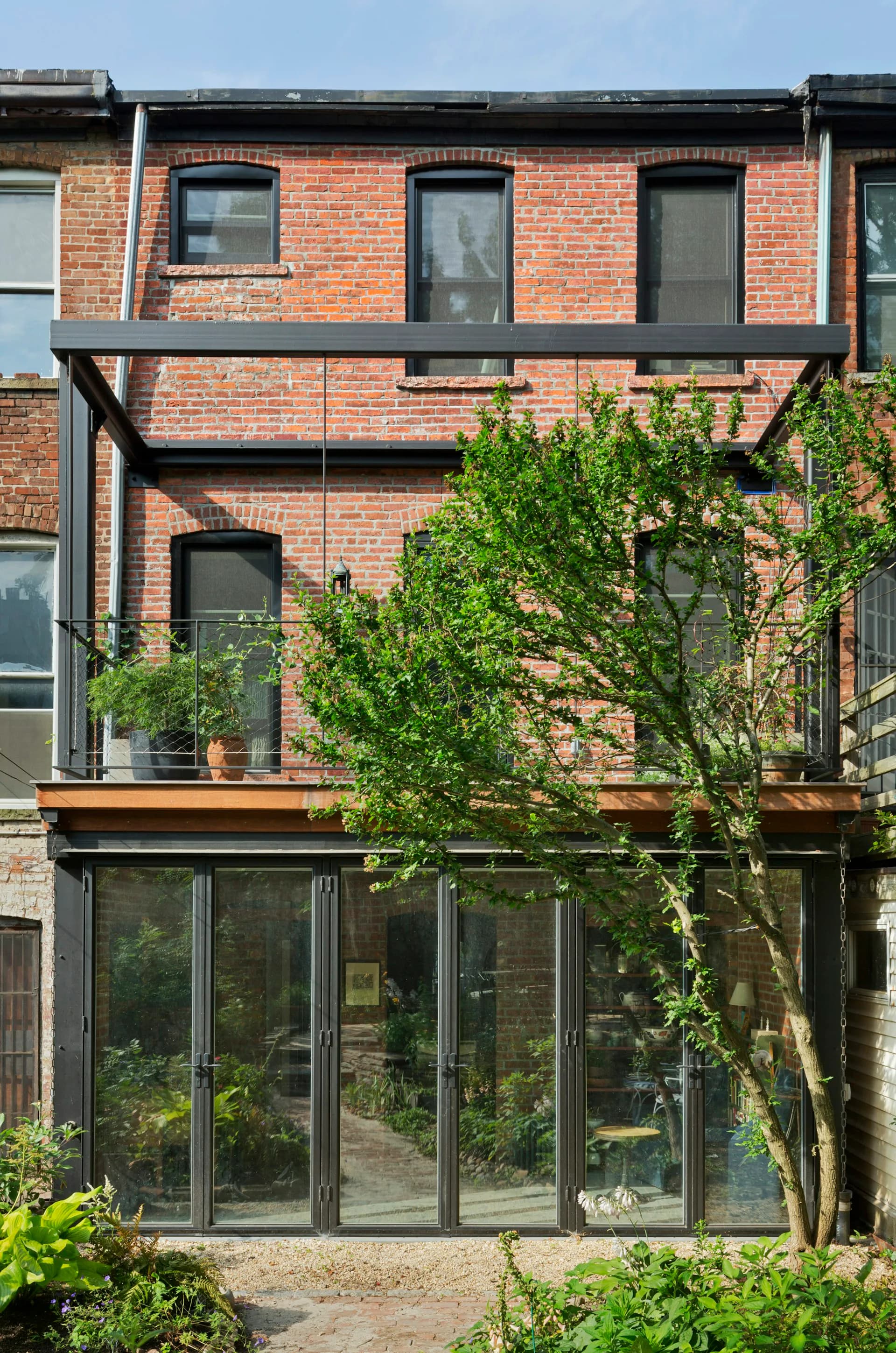 Contemporary brick facade with steel-framed windows and living green wall climbing across multiple levels, featuring modern glass doors and wooden deck elements by Gans studio.