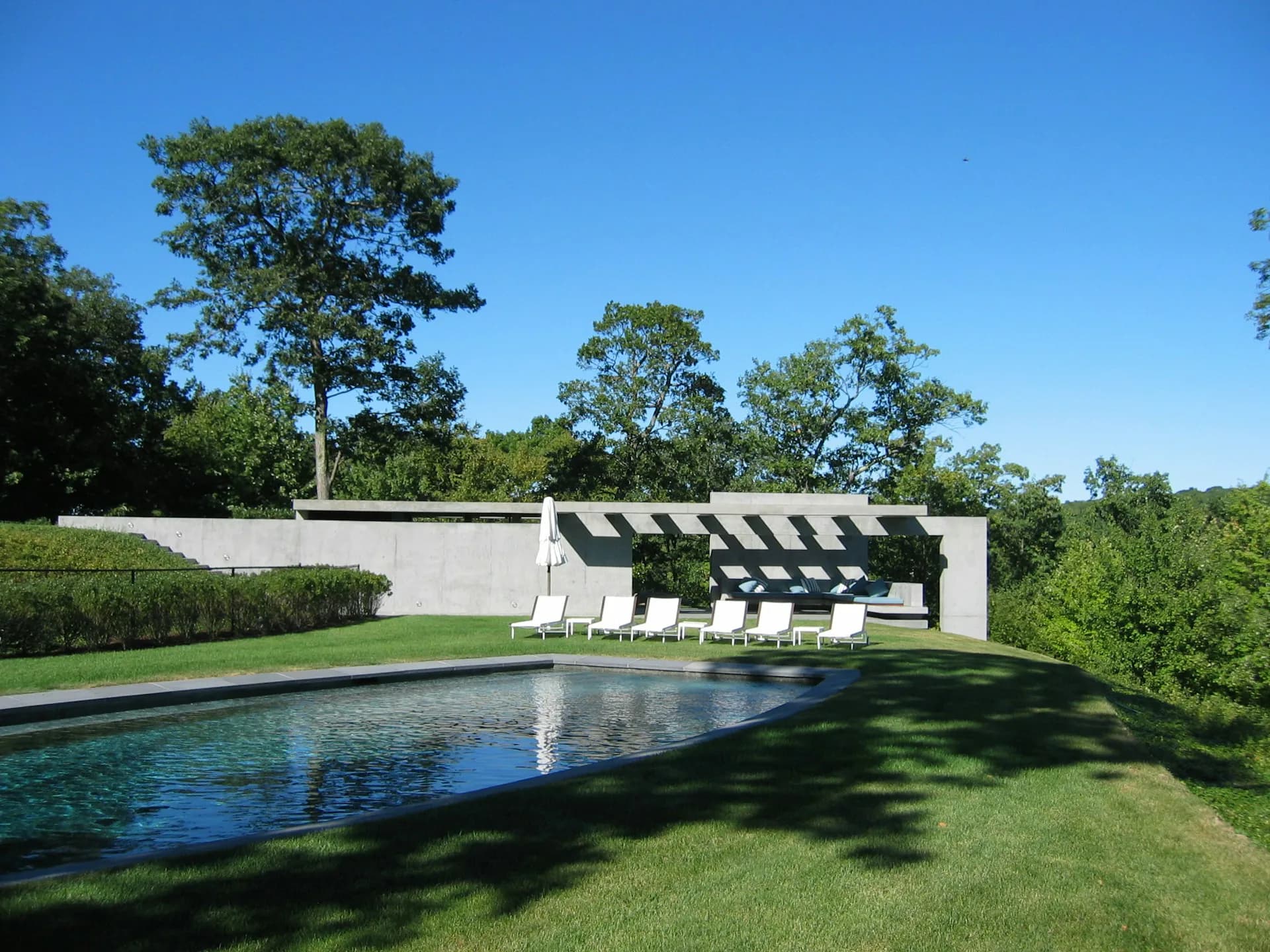 Modern poolside retreat featuring a rectangular swimming pool, white wall structure with black pergola, lounge chairs, manicured lawn, and wooded landscape backdrop by Gary Morgenroth Architect, LLC.