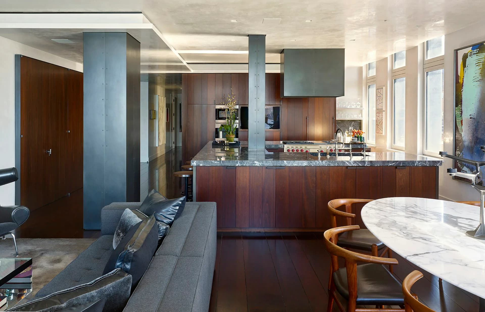 Modern open-plan apartment with dark wood kitchen island, marble dining table, gray seating, blue-gray columns, and abundant natural light by Gary Morgenroth Architect, LLC.