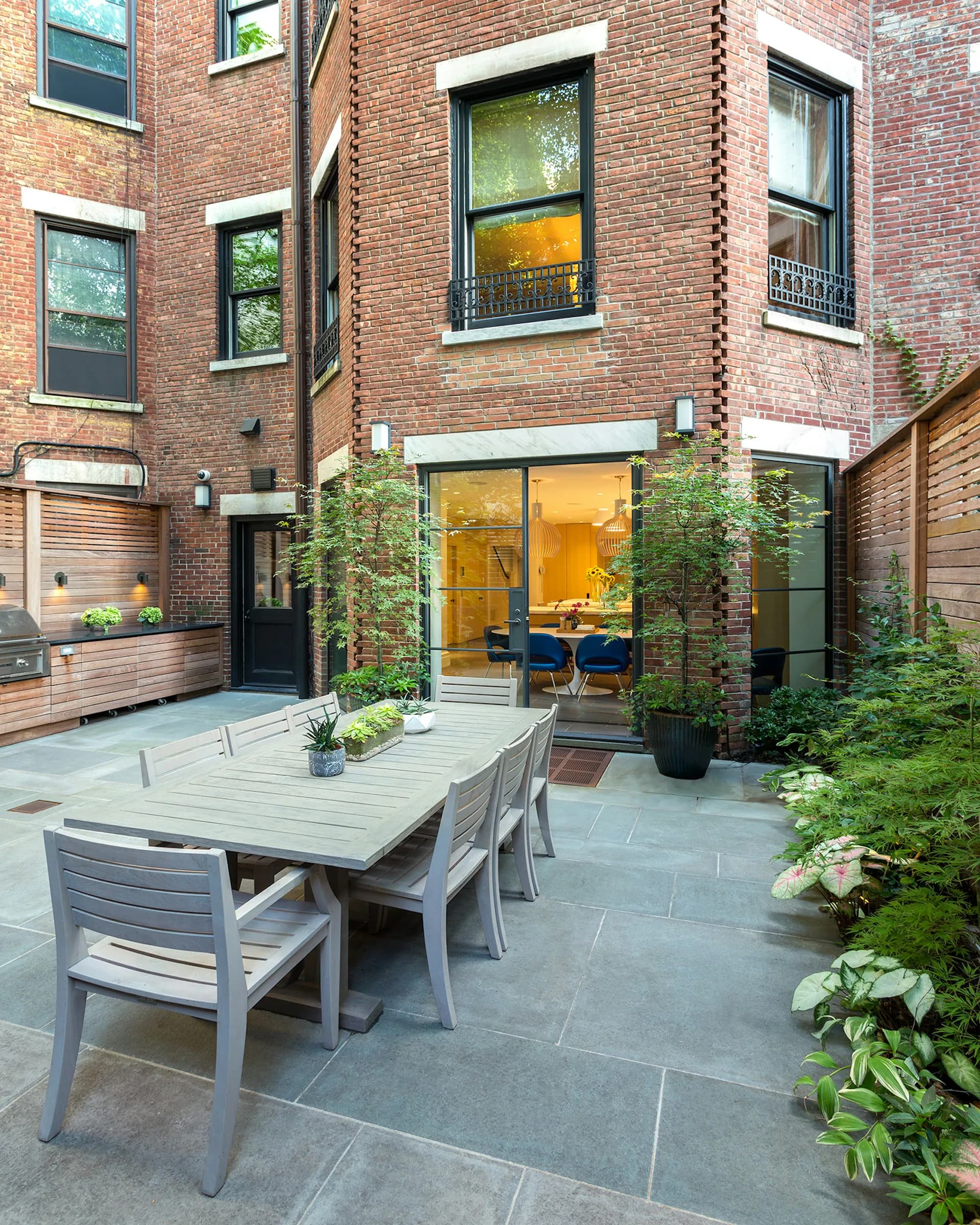 A contemporary brick courtyard features a dining table with gray chairs, lush green plantings, and large glass doors opening to an illuminated interior by Gary Morgenroth Architect, LLC.