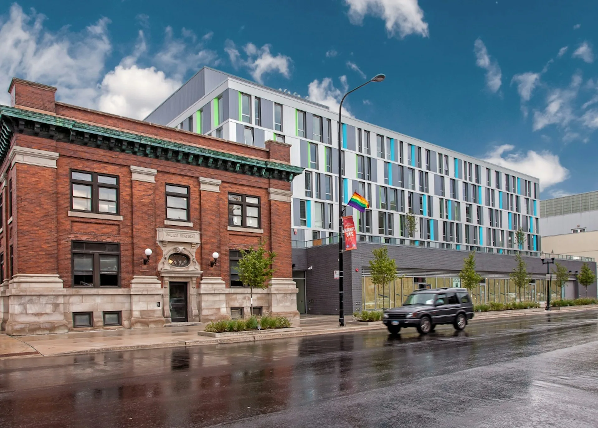 Mixed-use urban development with restored historic brick building and modern residential tower featuring green and blue facades by Gensler.