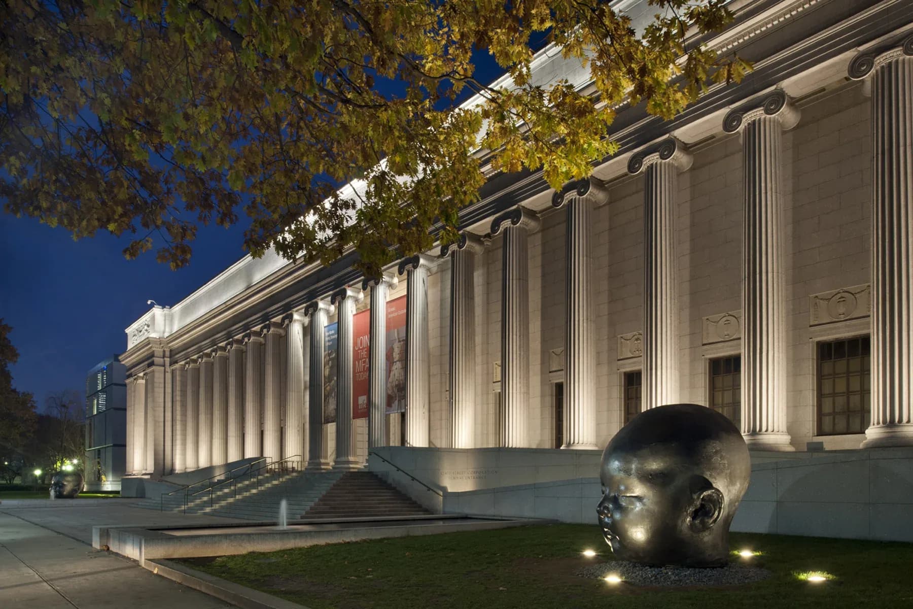 Classical museum facade with fluted columns illuminated at dusk, sculptural head in foreground, mature trees overhead, lighting design by George Sexton Associates.