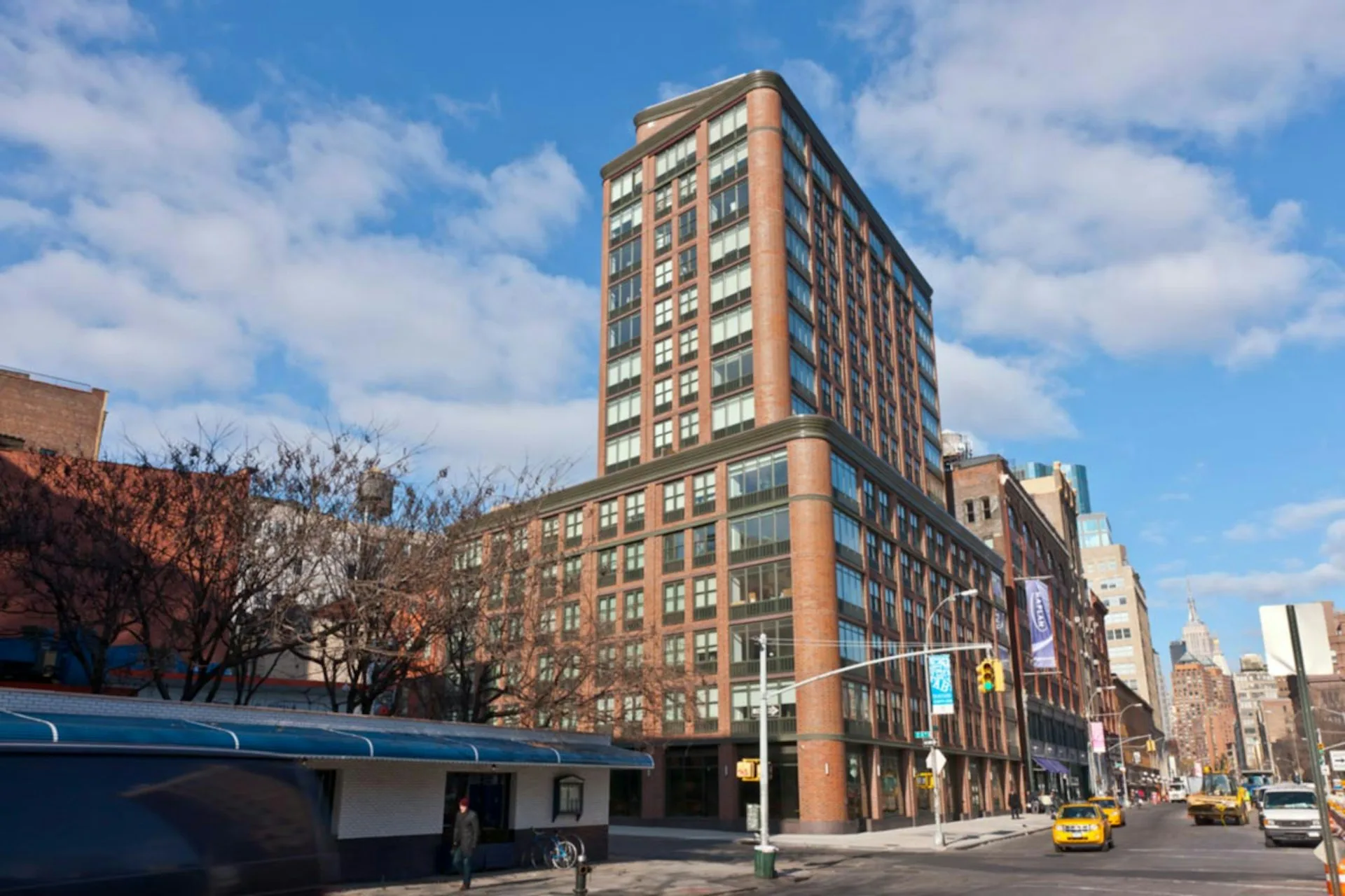 Corner view of a tan and brown brick residential building with regular window patterns, ground-floor retail, and yellow taxis on an urban street by Gerner Kronick + Valcarcel, Architects, DPC.