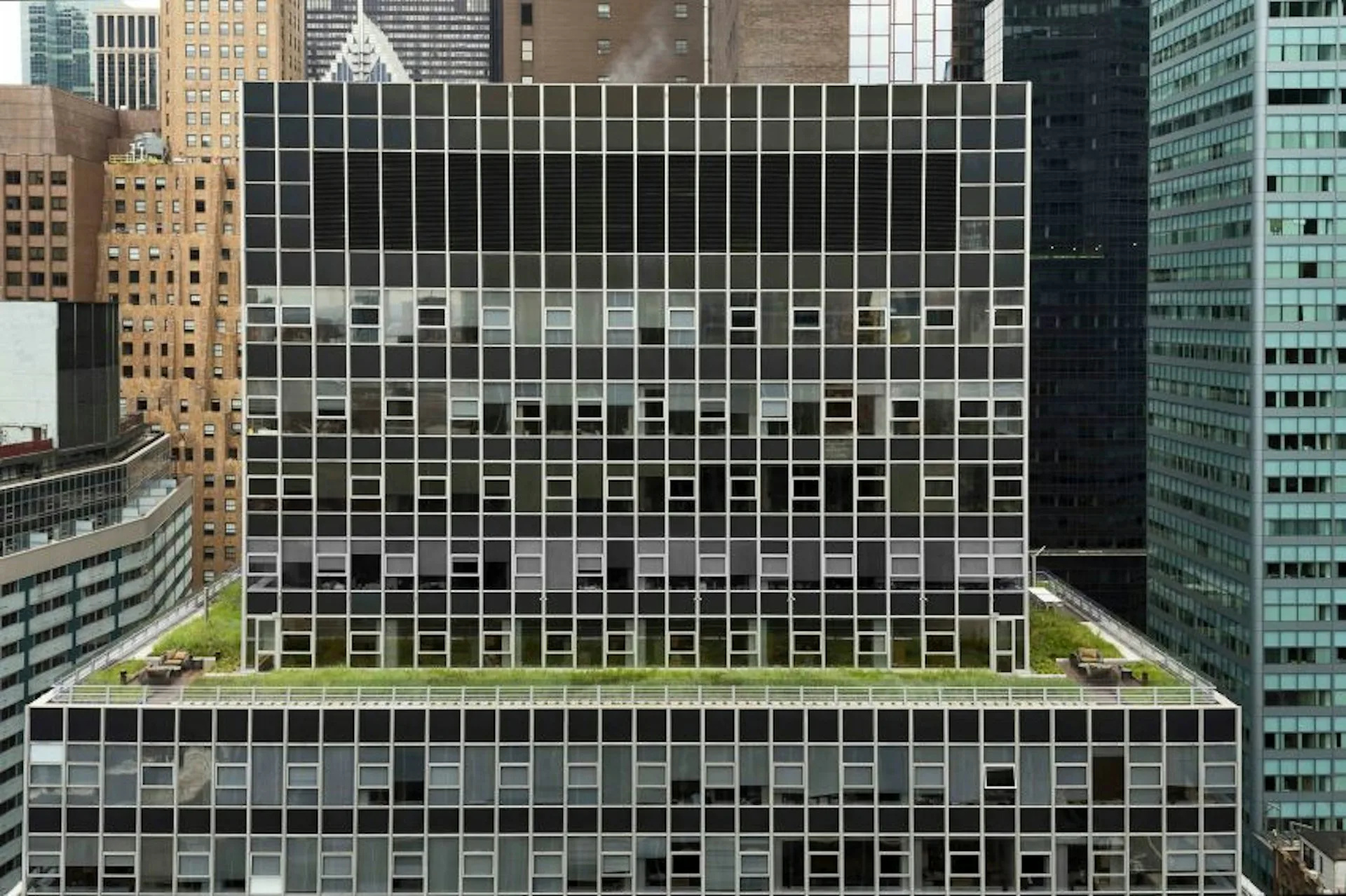 A modernist office building facade with a regular grid of black-framed windows and light-colored spandrel panels, surrounded by neighboring Manhattan skyscrapers, by Gertler & Wente Architects.