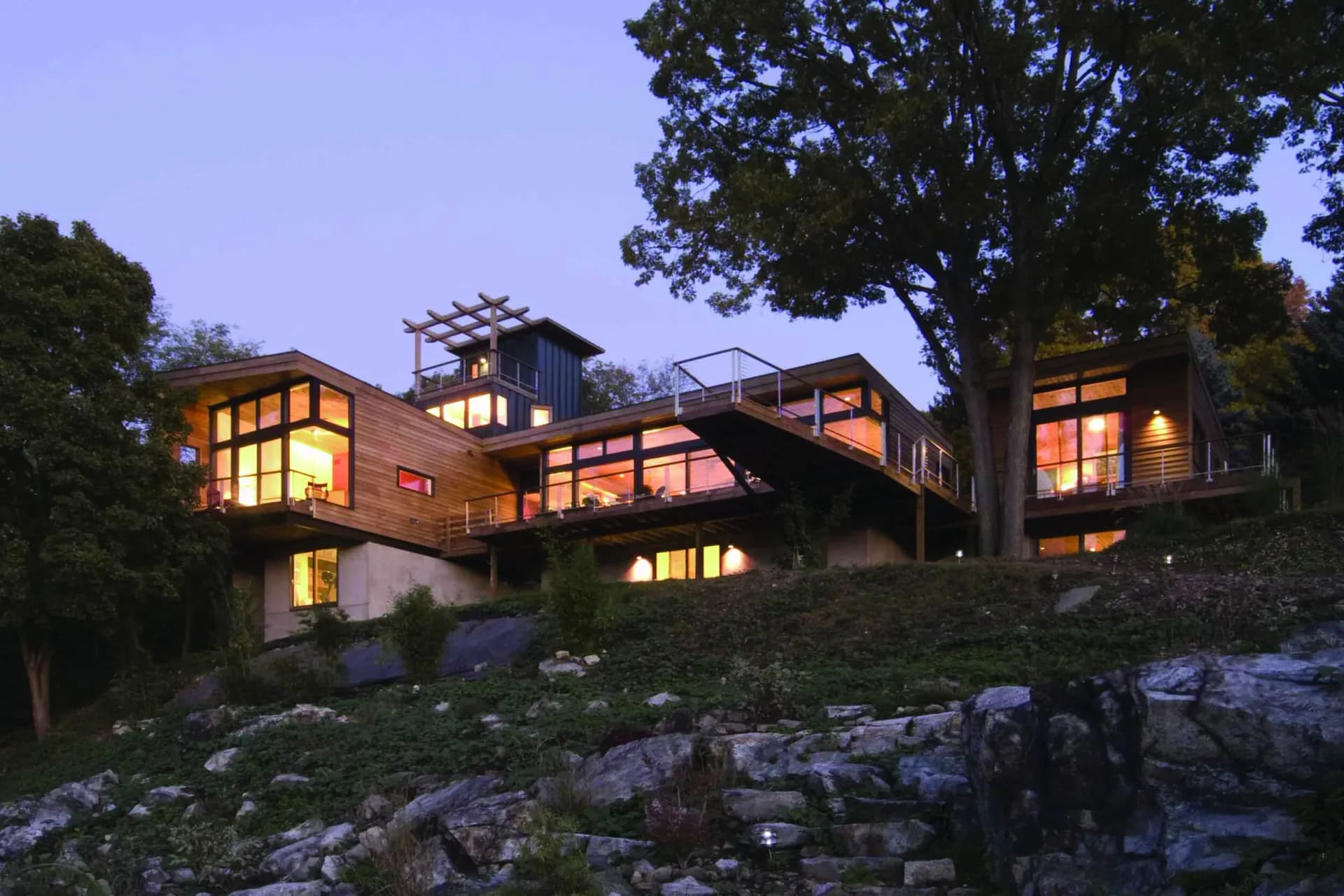 Modern residential architecture photographed at dusk with warm interior lighting, featuring multi-level wooden and stone construction nestled among mature trees by Gertler & Wente Architects.