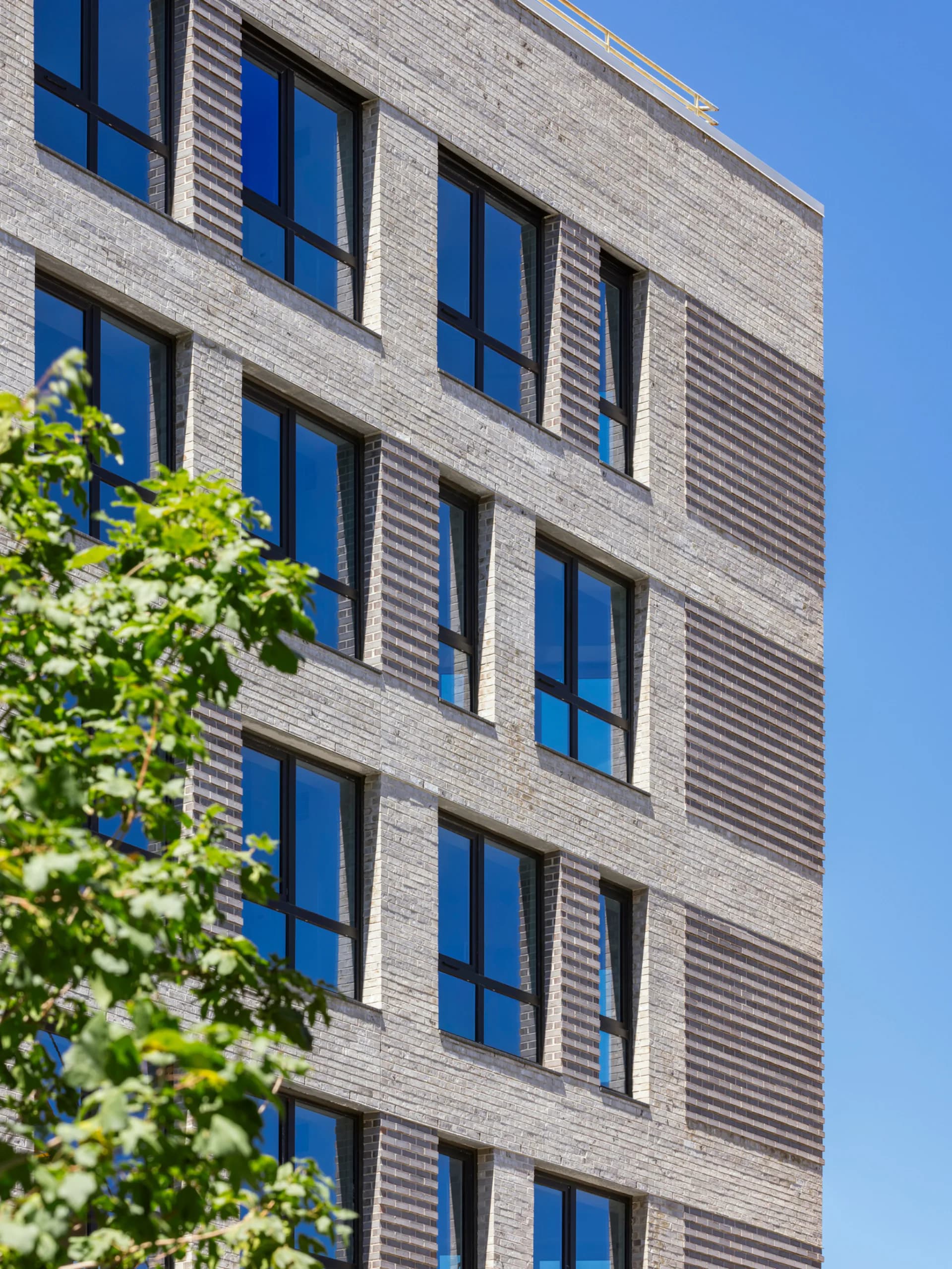 Contemporary brick building facade with five stories of large blue-framed windows and horizontal metal louvers, clear sky background, designed by GF55 Architects.