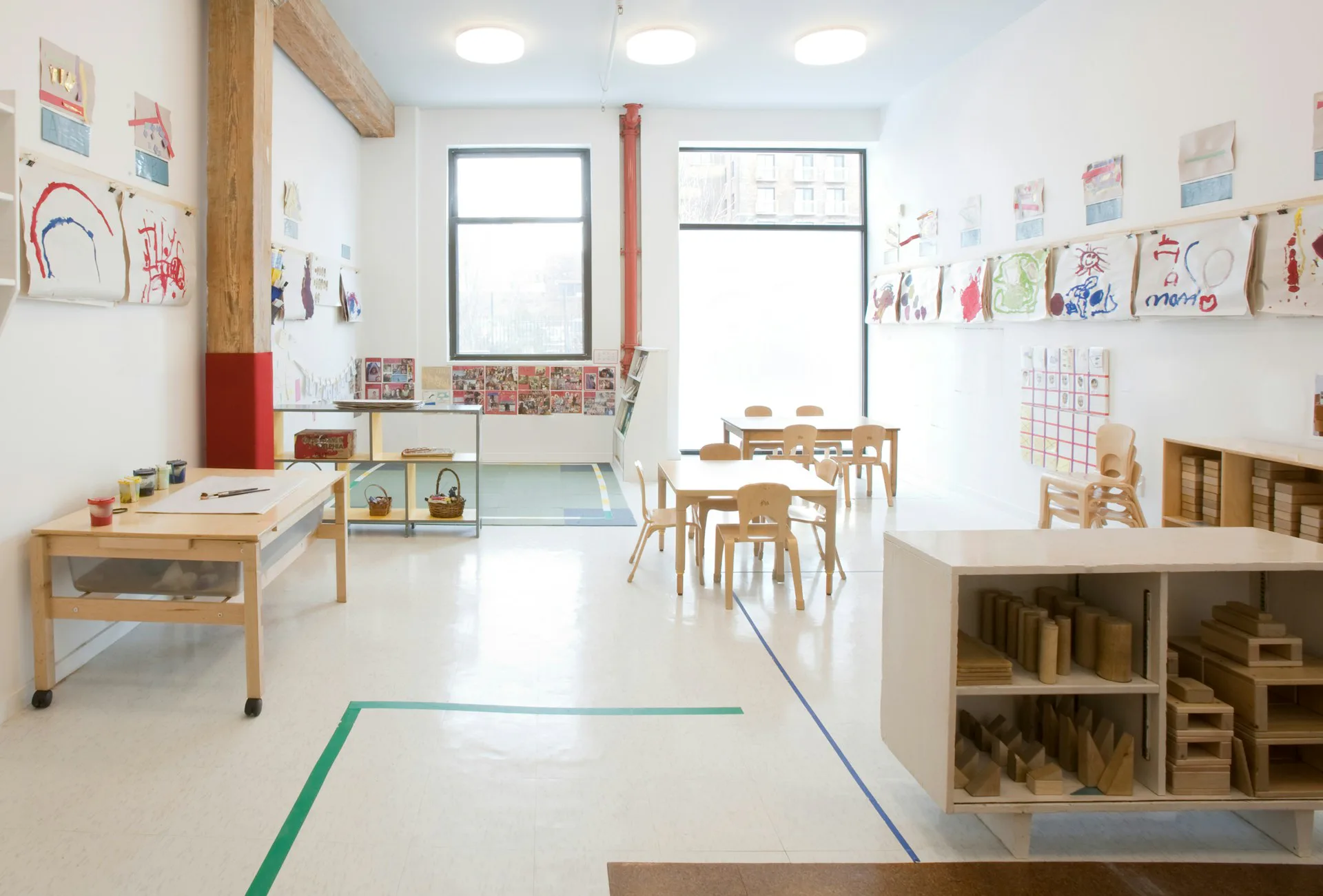 Bright Montessori classroom with light wood furniture, colorful children's artwork on walls, green floor tape dividers, and natural light from large windows, by Glickman Schlesinger Architects.