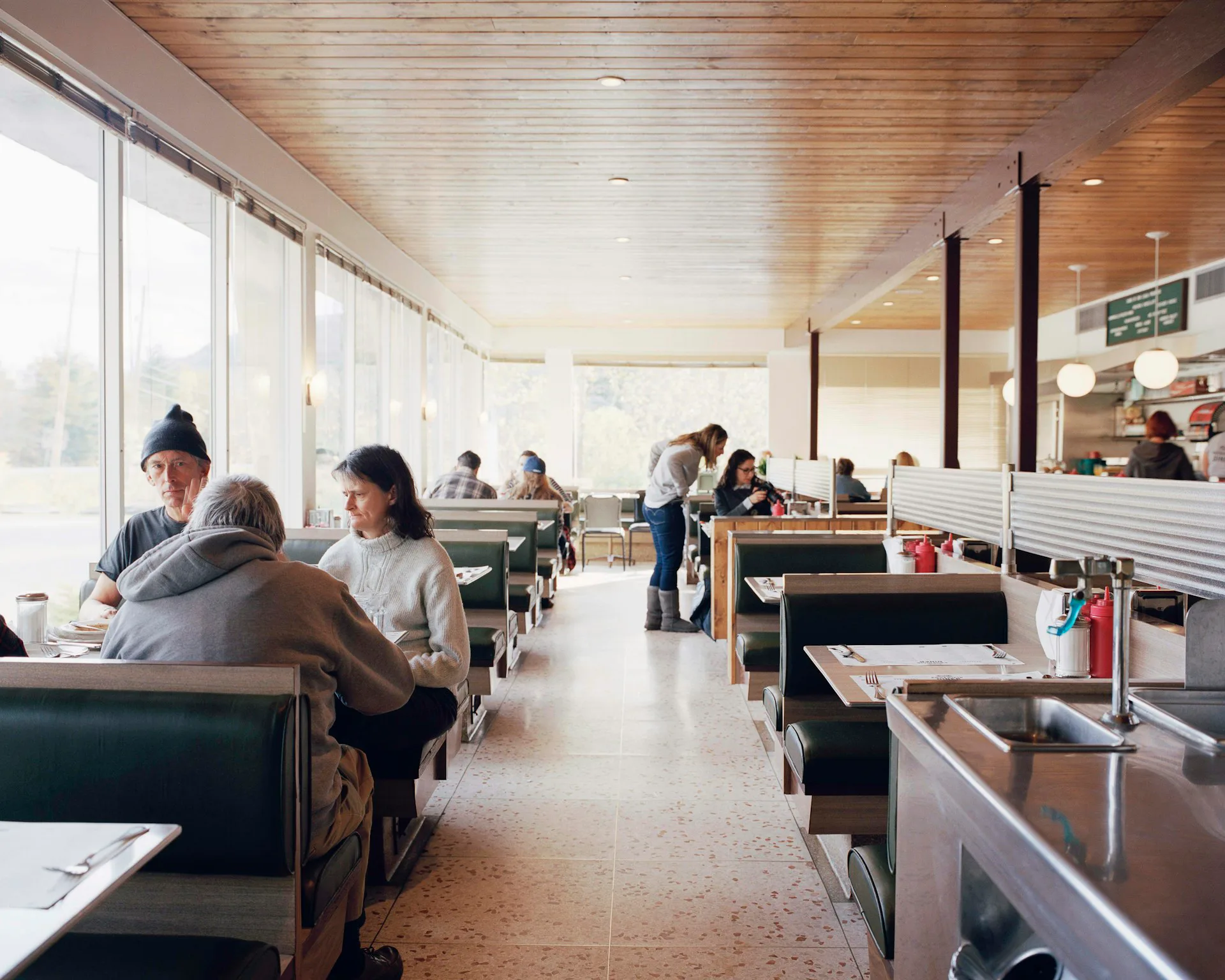 A bright diner interior with wood ceiling, booth seating along windows, stainless steel counter, and warm lighting by Glickman Schlesinger Architects.