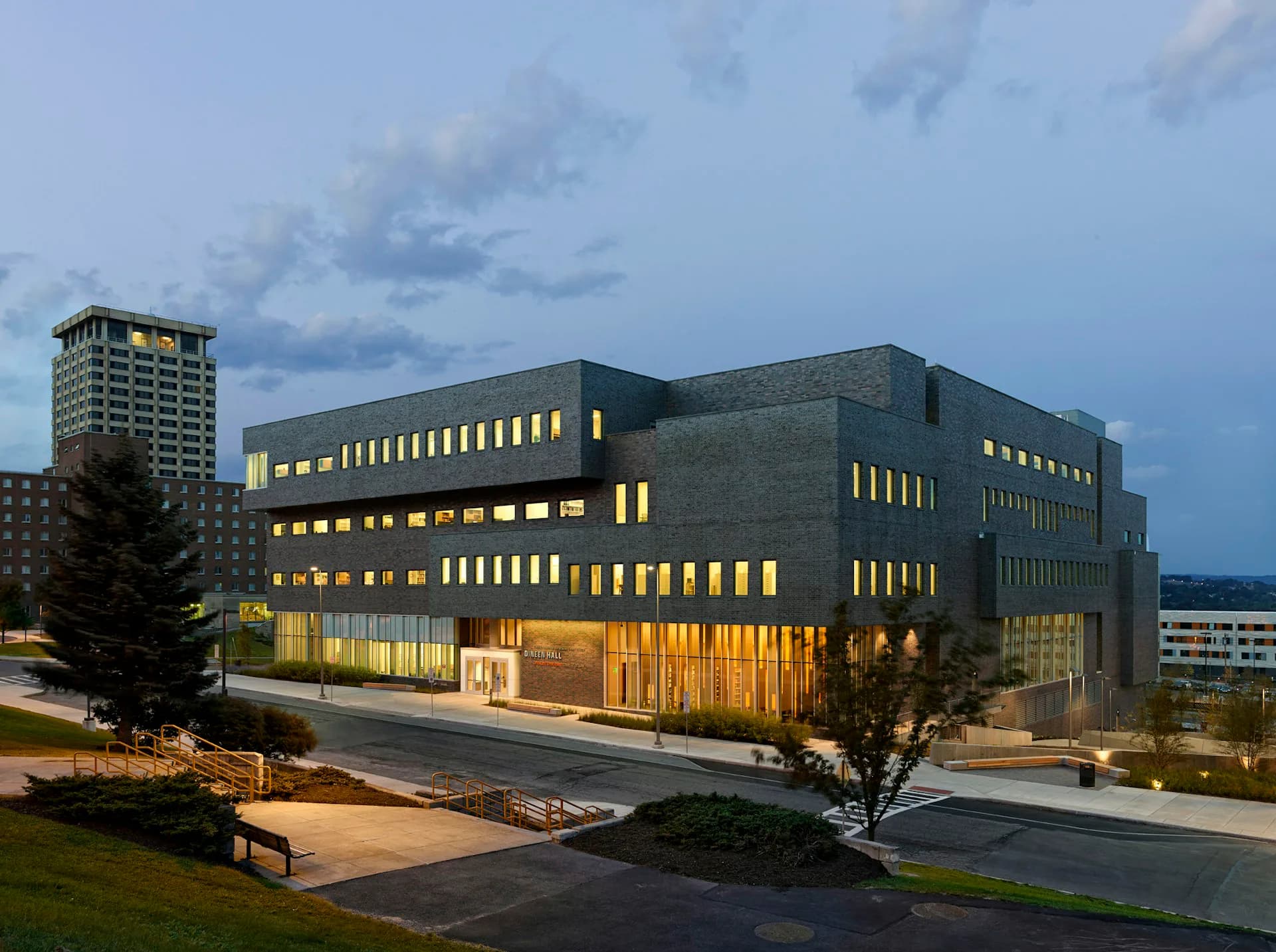 Modern institutional building with dark brick facade and illuminated golden-yellow window frames photographed at dusk, surrounded by landscaping and plaza by Gluckman Tang Architects.