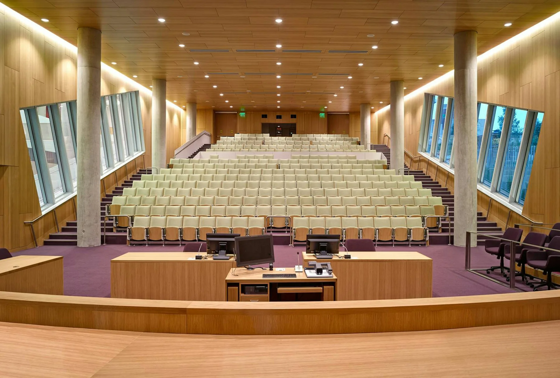 Modern auditorium interior with cream-colored tiered seating, warm wood paneling, recessed lighting, and purple-carpeted stage platform designed by Gluckman Tang Architects.
