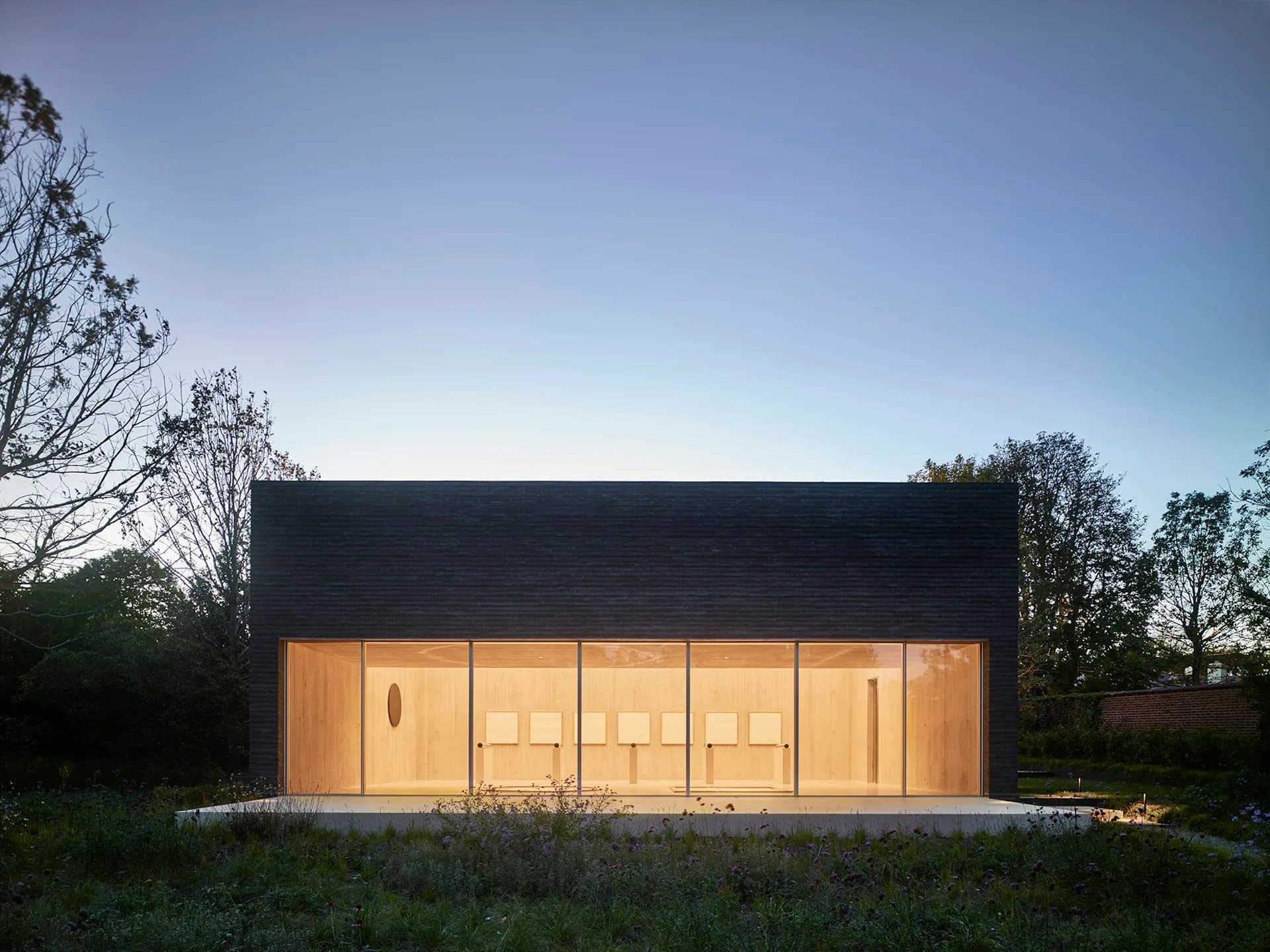 Modern pavilion with black roof and warm timber-clad facade featuring large glazed doors, illuminated at dusk against natural landscape by Gluckman Tang Architects.
