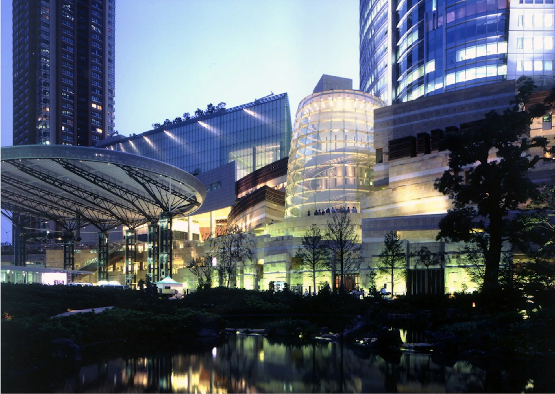 Illuminated modern architectural complex at dusk featuring a cylindrical glass tower and geometric structures reflected in water, by Gluckman Tang Architects.