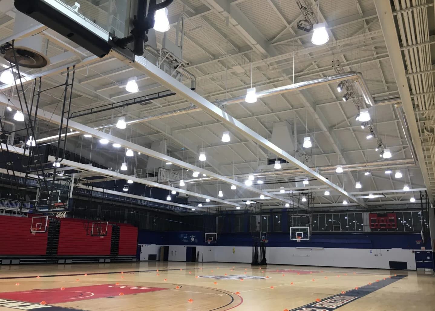 Modern gymnasium interior with red and navy blue hardwood court, LED ceiling lighting, and retractable bleachers by Goldstick Studio.