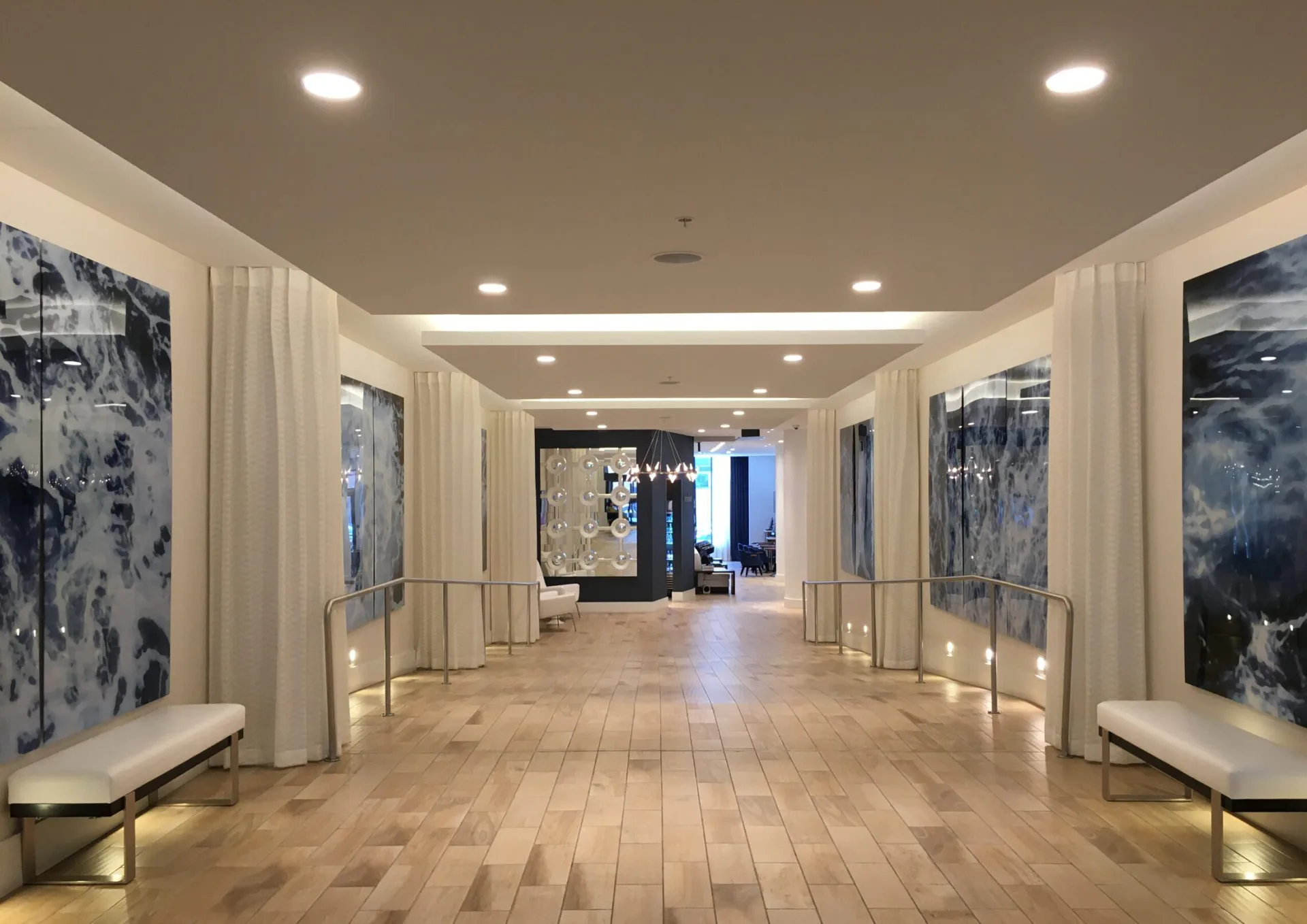 A gallery corridor with cream-colored curtains, wood flooring, recessed lighting, and large artworks displayed on side walls, designed by Goldstick Studio.