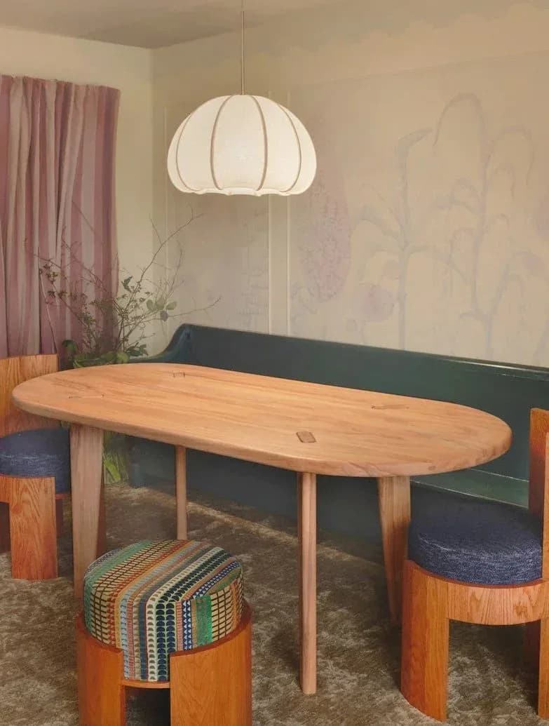 Warm wood dining table with rounded ends paired with sculptural wooden chairs and a woven pouf, lit by a organic pendant fixture, by Good Chair Co.