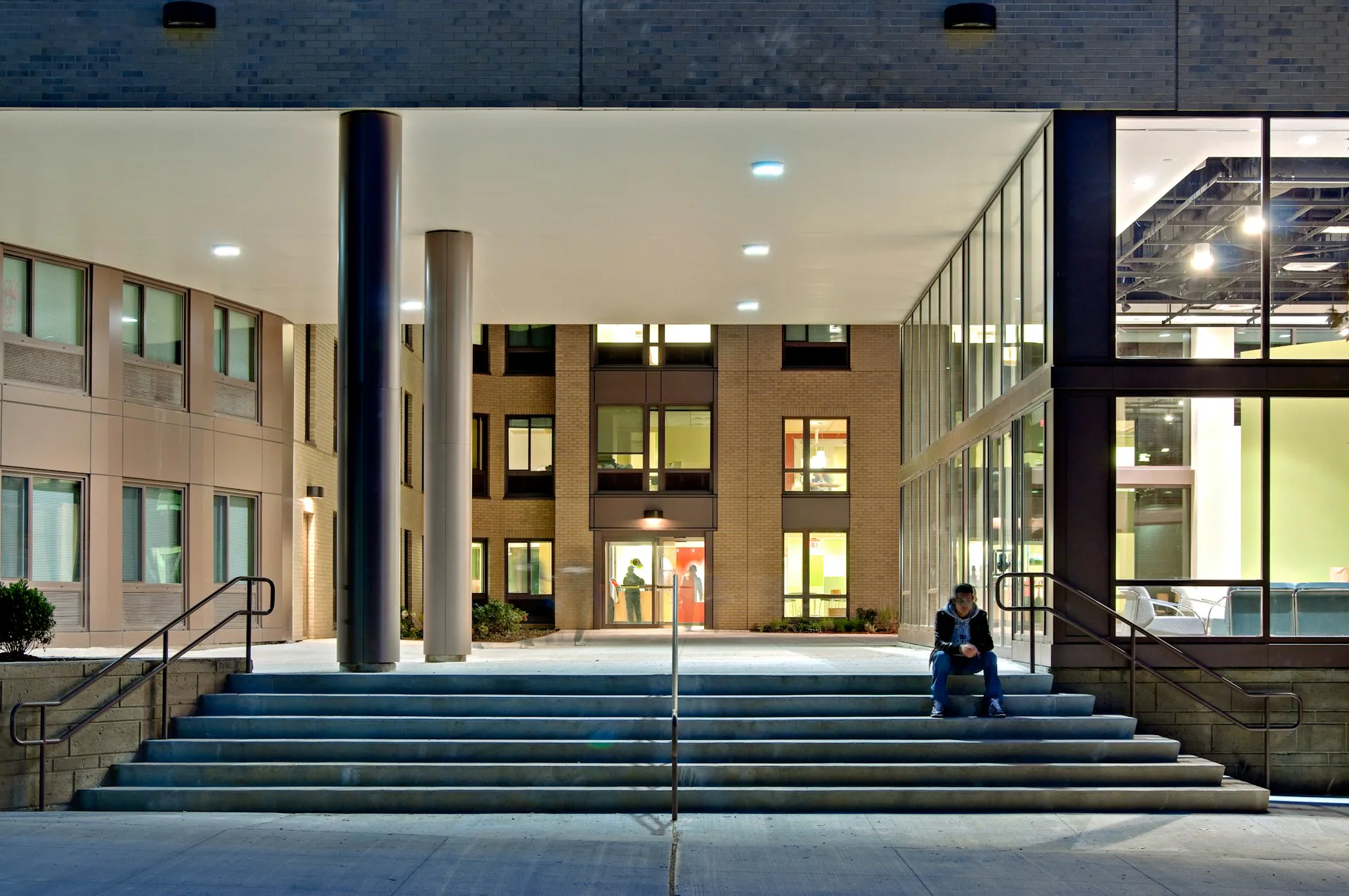 Modern building entrance with wide blue-gray steps, glass doors, and illuminated interior spaces with warm lighting, designed by Goshow Architects.