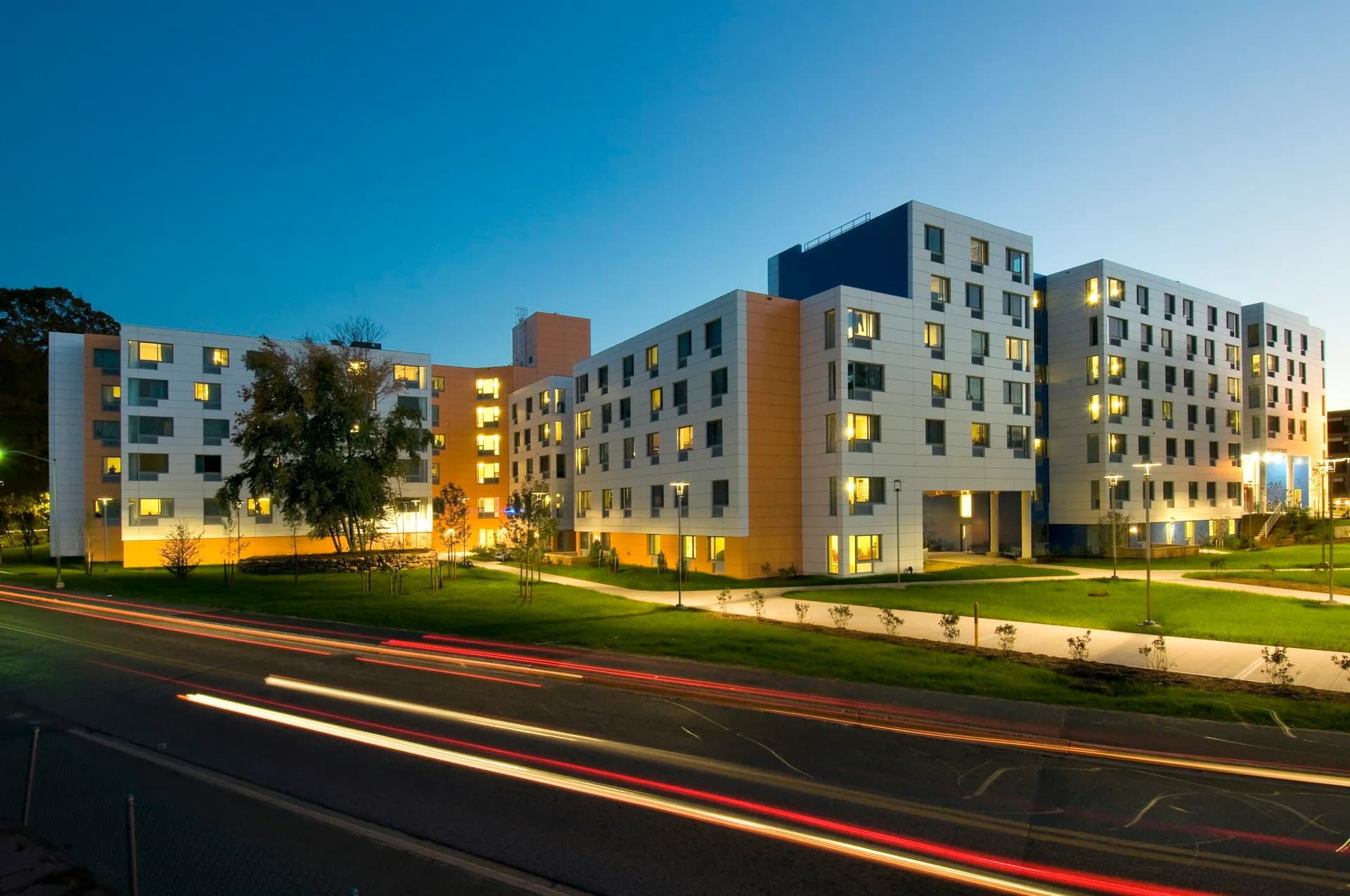 Modern residential apartment complex illuminated at dusk with yellow-lit windows, clean geometric facades in blue and tan tones, manicured lawn, and street traffic by Goshow Architects.