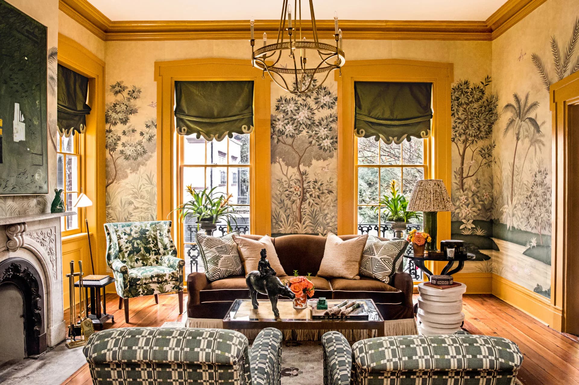 Mustard-yellow sitting room with botanical wallpaper, brass chandelier, dark sofa, patterned armchairs, fireplace, and tall windows dressed with Roman shades by Gracie.
