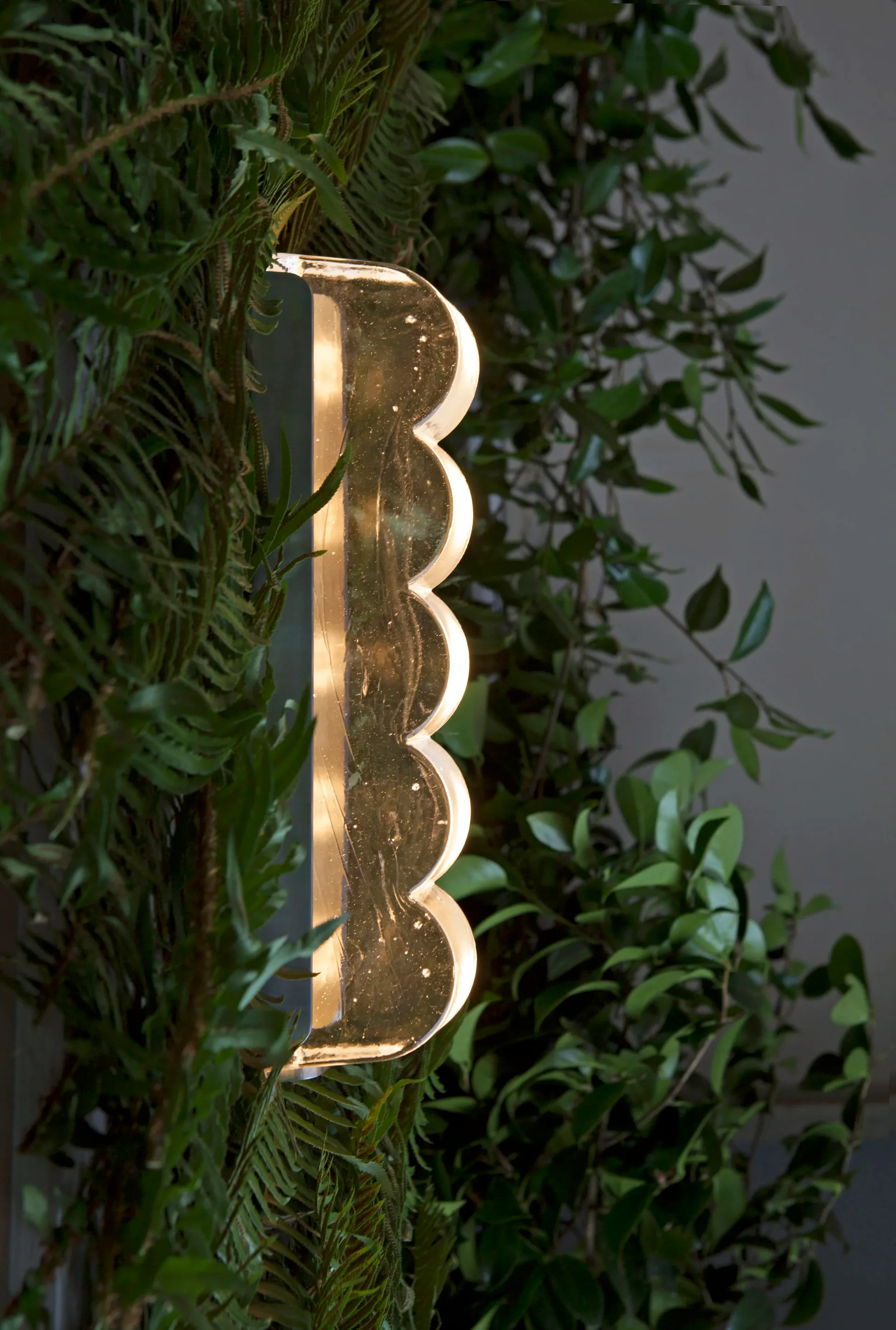 Golden wavy-edged vertical light fixture mounted on foliage-covered wall, creating warm illumination, by Grain Design.