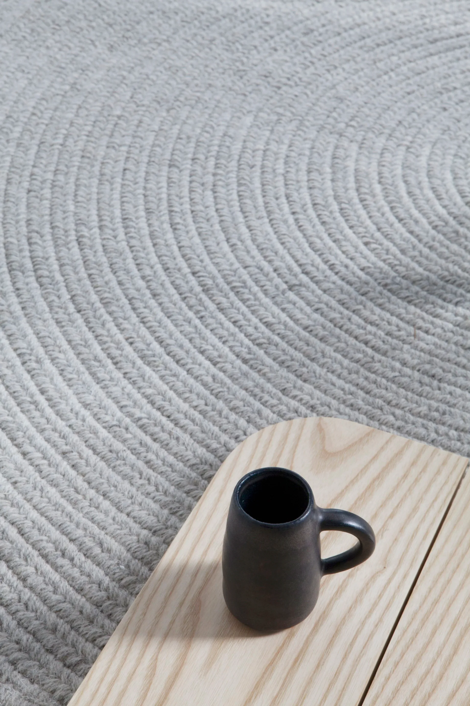 Minimalist interior detail showing a black ceramic mug on light wood surface against textured gray wall by Grain Design.