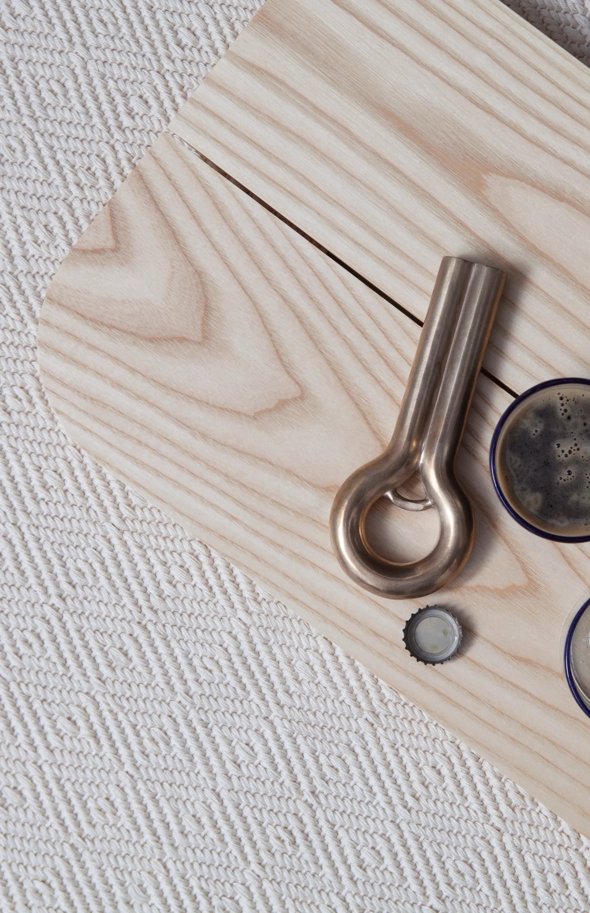 Minimalist wooden surface with stainless steel bottle opener and beer caps arranged as a styled still life by Grain Design.