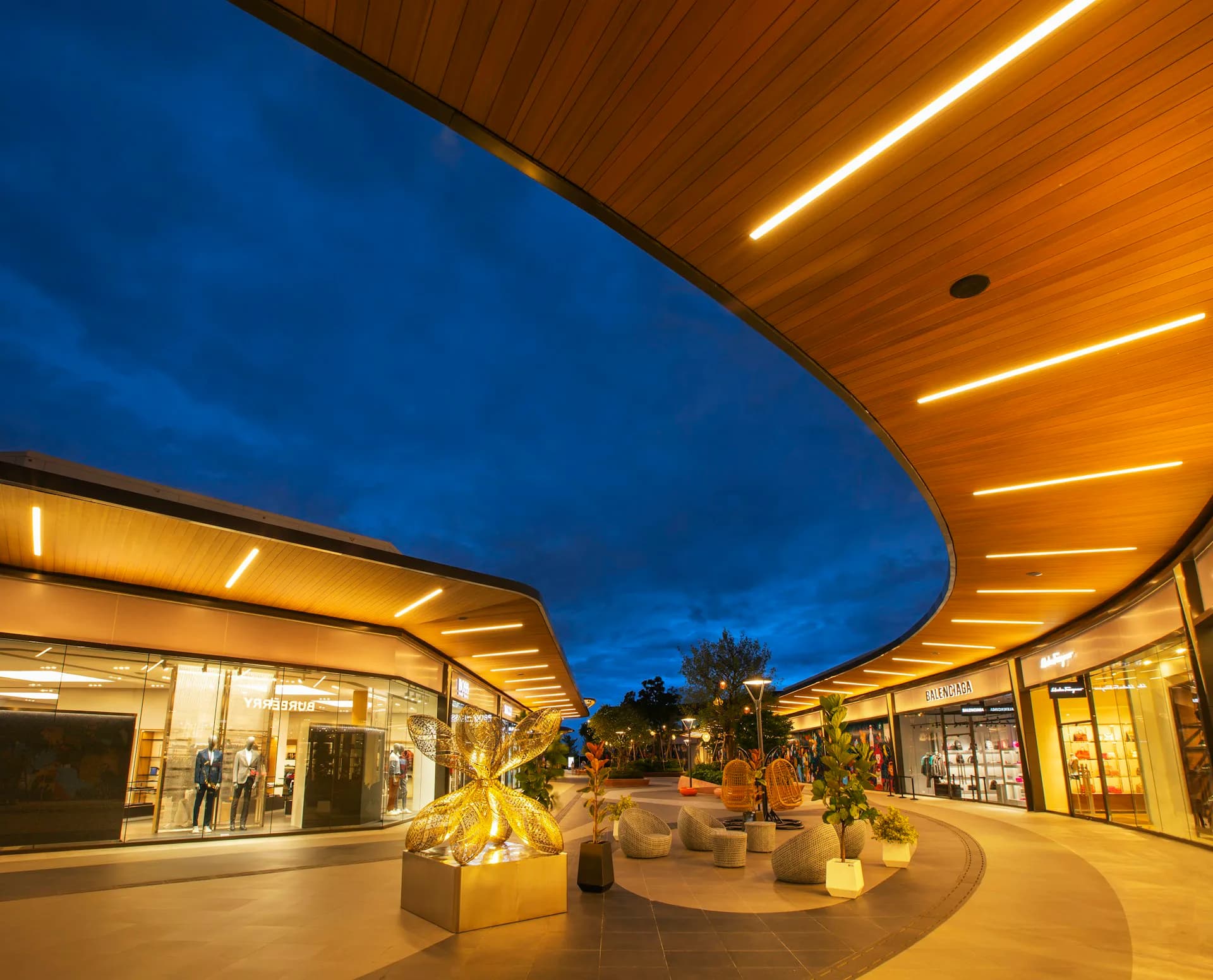 Evening view of a modern curved shopping pavilion with warm wood ceilings and glowing storefronts beneath a twilight blue sky by Grimshaw Architects.