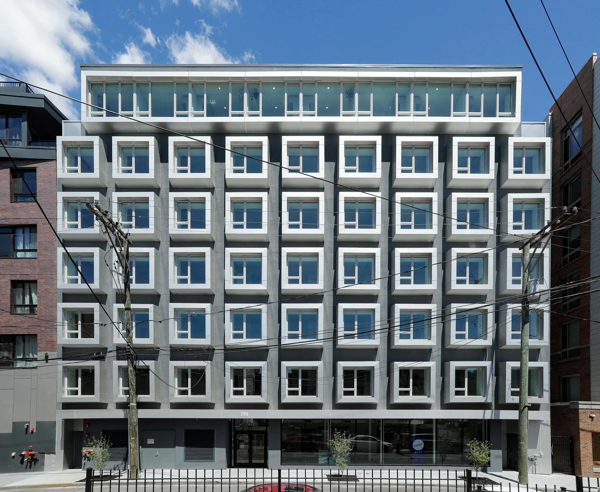 Modern multi-story residential building with teal-blue facade, repetitive grid of white-framed windows, ground-floor commercial spaces, and clean contemporary design by GRO Architects.