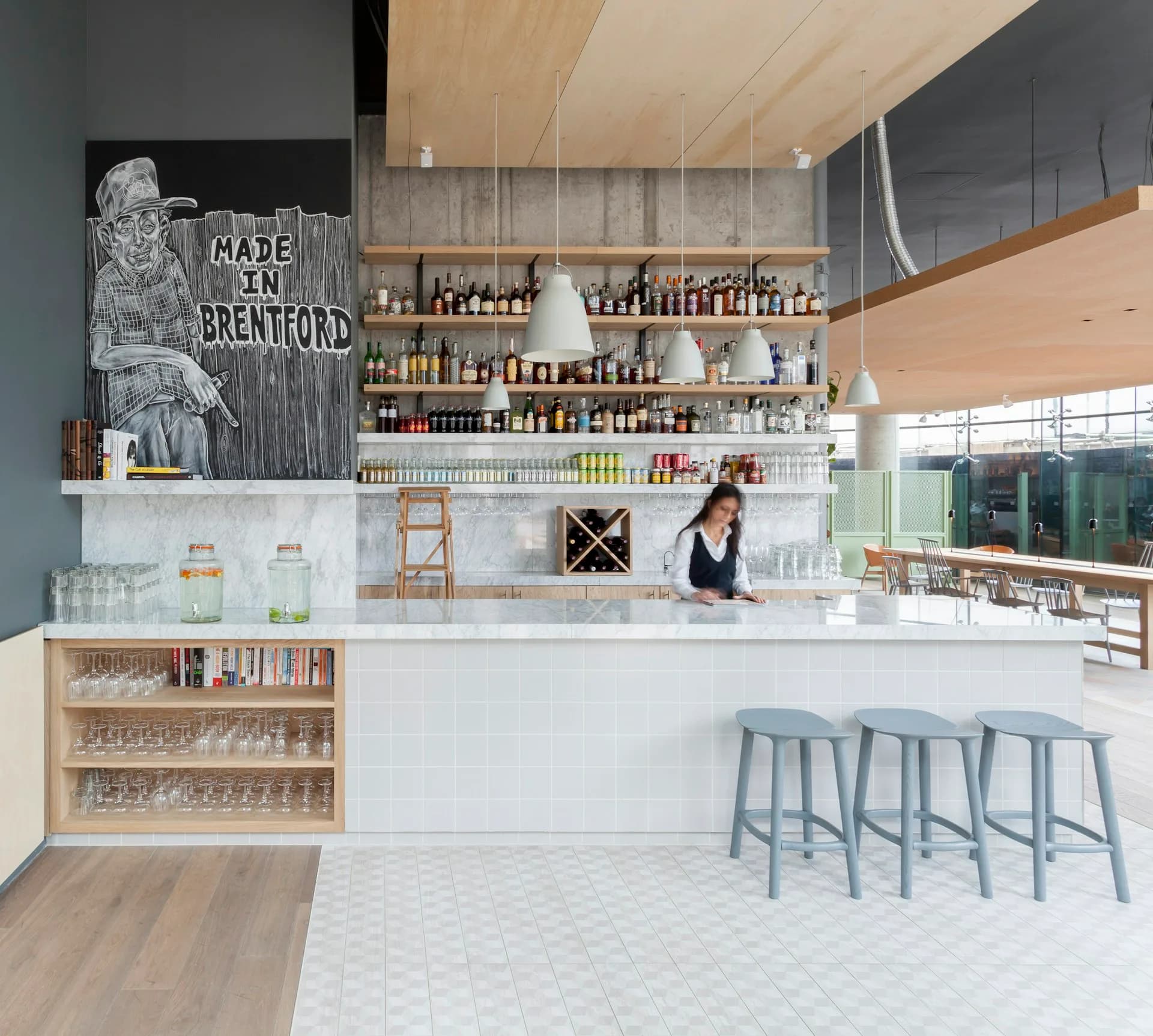 Modern apothecary or natural products retail interior with white counter, wooden ceiling, shelving displaying amber glass bottles, and blue bar stools, designed by Grzywinski+Pons.
