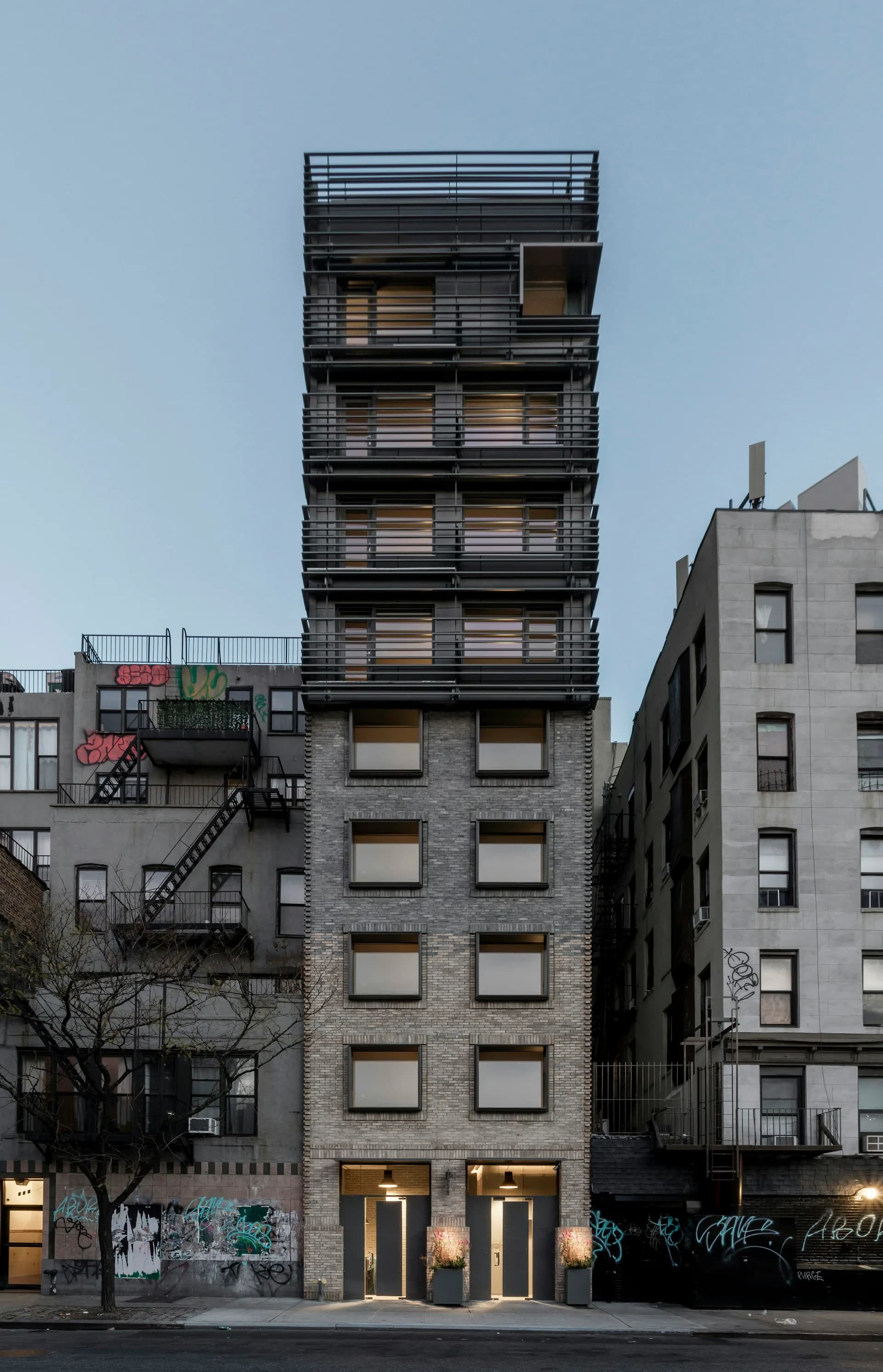 Tall residential building with dark horizontal louvers above brick base, street-level storefront with golden lighting, surrounded by older urban buildings, designed by Grzywinski+Pons.