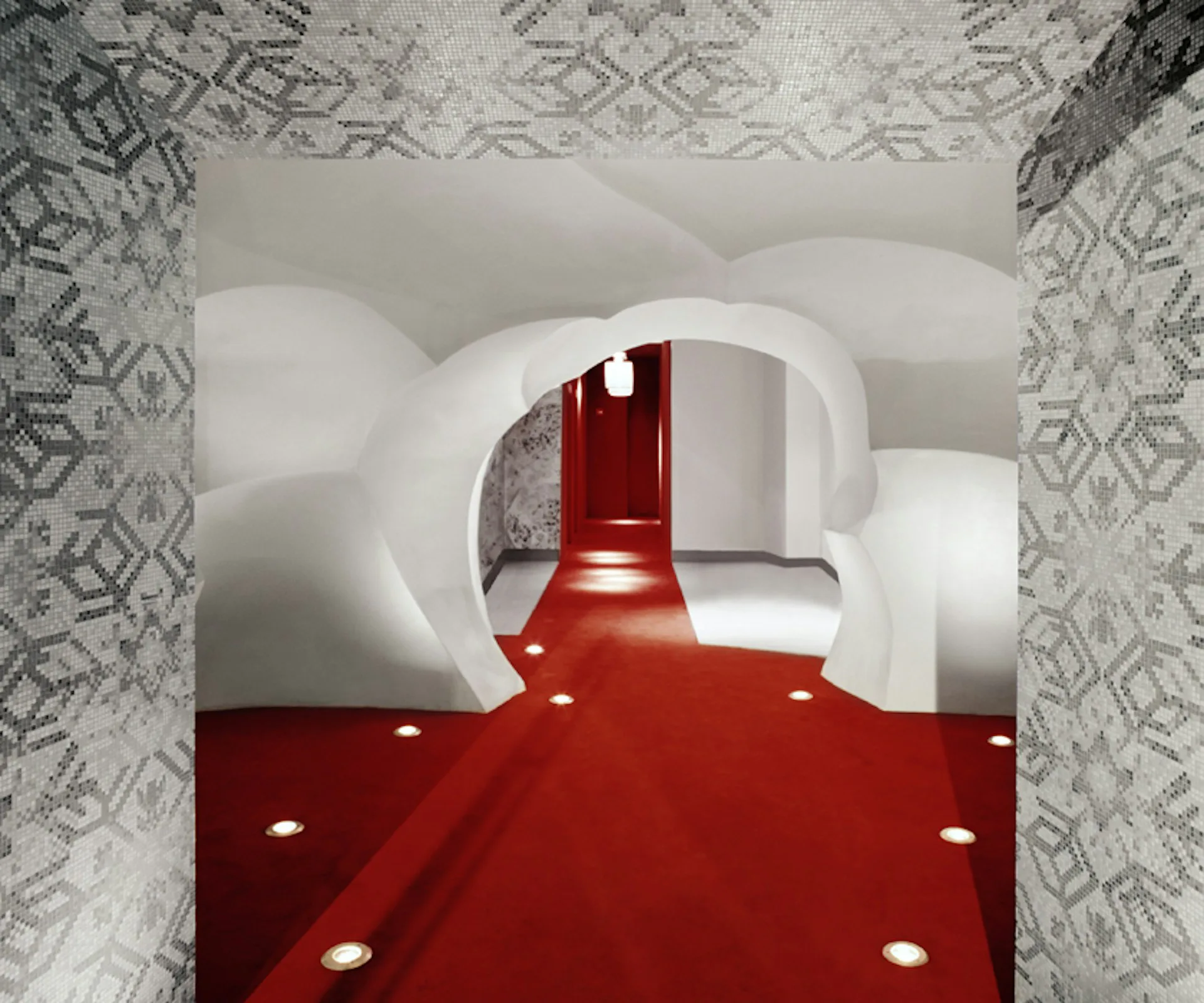 A curved white interior hallway with dramatic red flooring and recessed lighting, featuring geometric patterned wallpaper framing the view by Grzywinski+Pons.