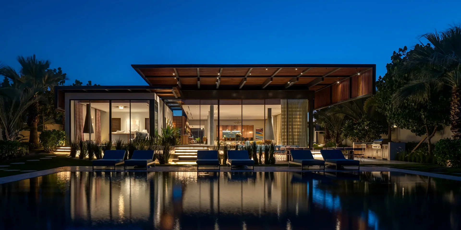 Modern waterfront residence with glass walls, wooden pergola, and reflecting pool at dusk, illuminated interiors visible, by Guerin Glass Architects.