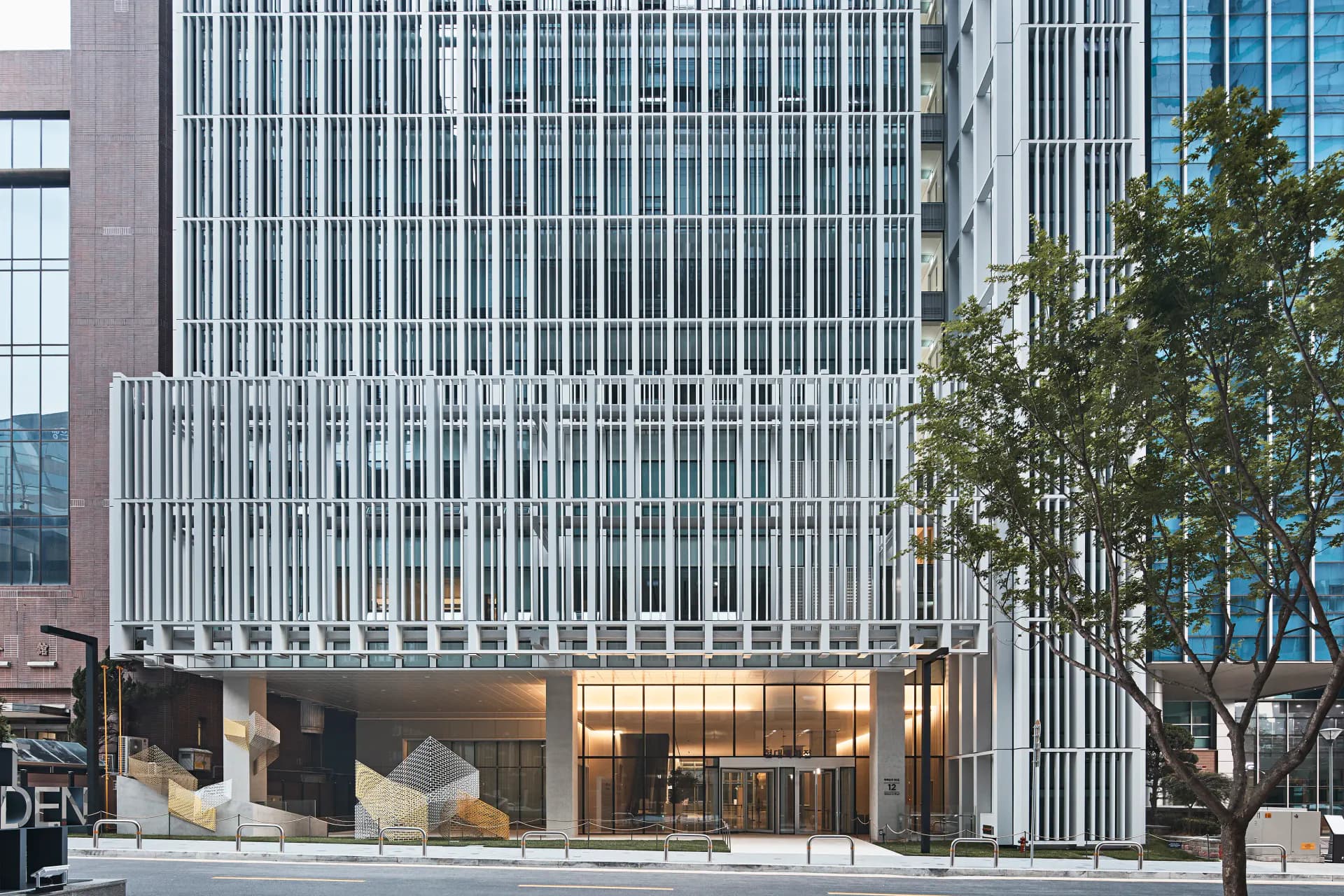 Modern commercial building facade with white vertical ribbed cladding, ground-floor glazing, and integrated landscaping by H ARCHITECTURE.
