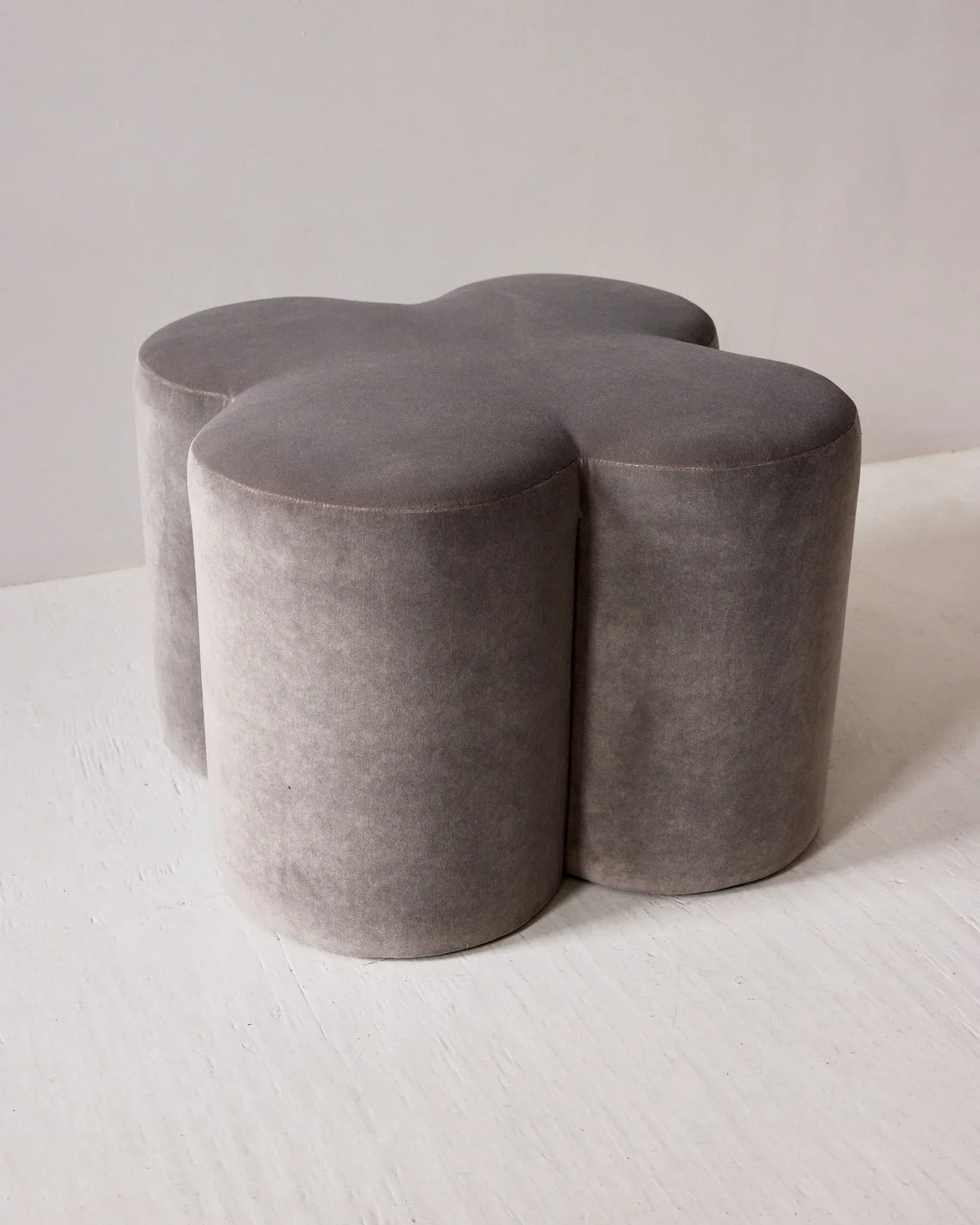 Four cylindrical gray upholstered ottoman poufs arranged in a clustered formation on a white floor, showcasing contemporary furniture design by H. Bigeleisen Studio.