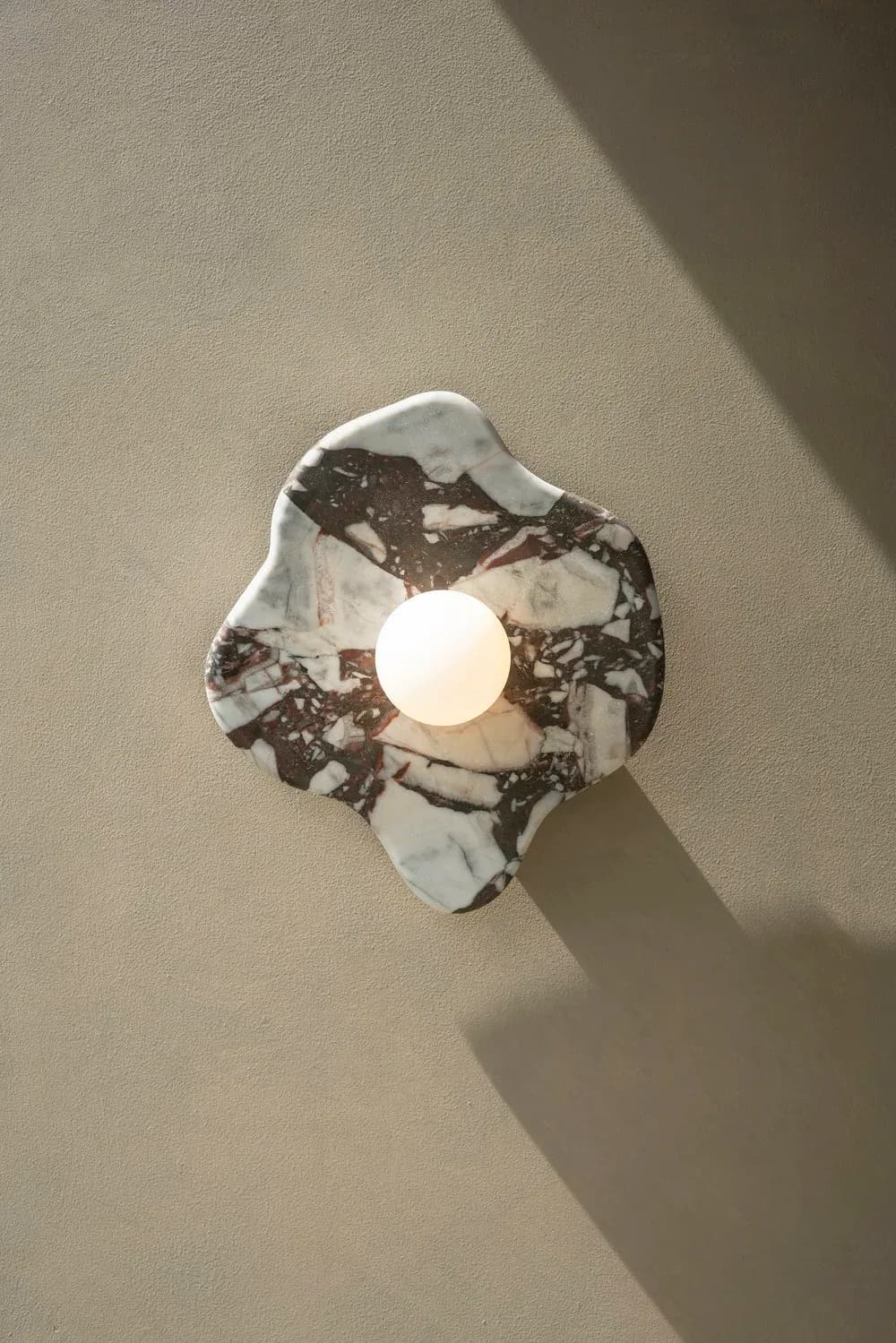 Contemporary wall-mounted light fixture with organic marble form and cream-colored sphere, casting dramatic shadows on neutral wall by H. Bigeleisen Studio.