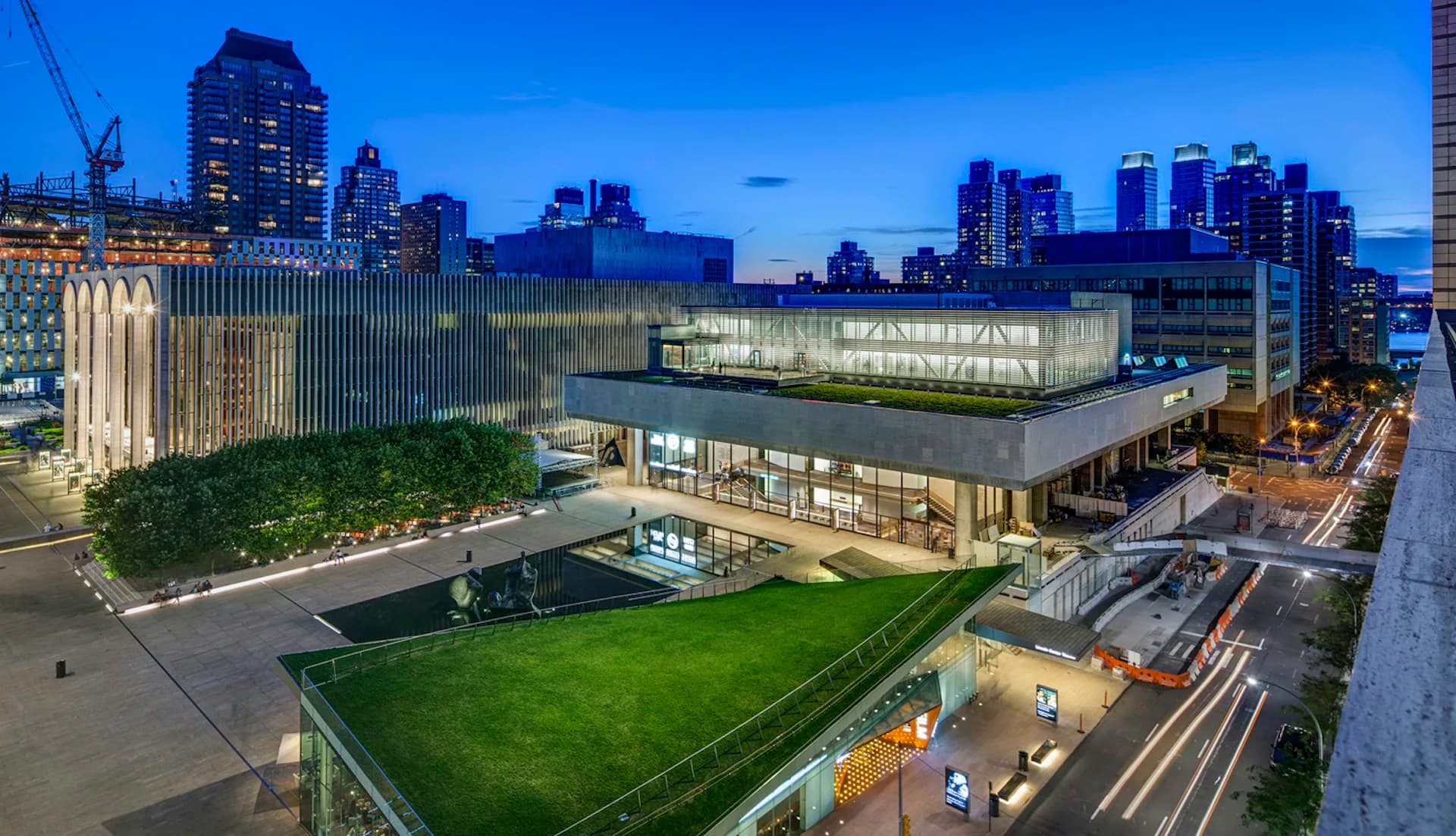 Modern institutional building with illuminated glass facade and rooftop green space overlooking Manhattan skyline at dusk, designed by H3 Hardy Collaboration Architecture, LLC.