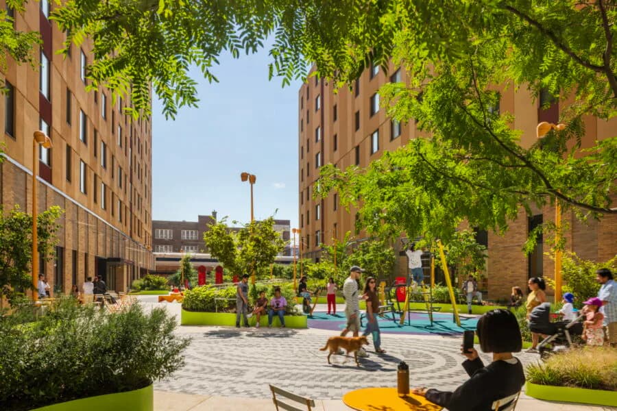 Sunlit courtyard with brick residential buildings, green trees, seating areas, and people gathered in the plaza designed by Handel Architects.