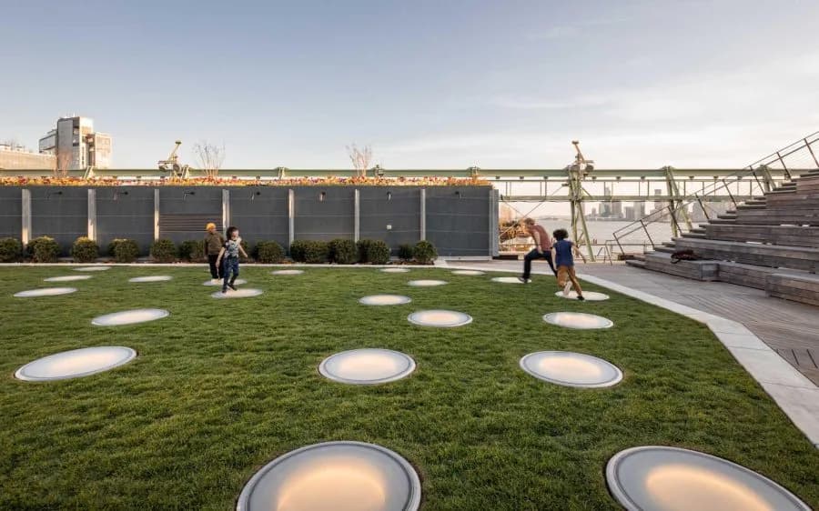 Rooftop green space with illuminated circular ground features and grass lawn, modern industrial backdrop by Handel Architects.