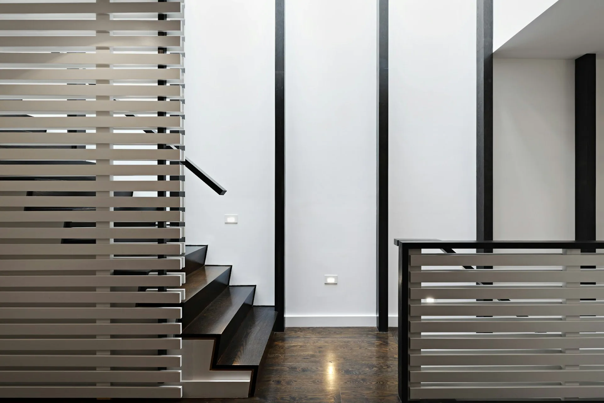 Modern staircase with horizontal slatted metal railings in a minimalist interior featuring white walls, dark flooring, and geometric architectural detailing by Hanson Architecture.